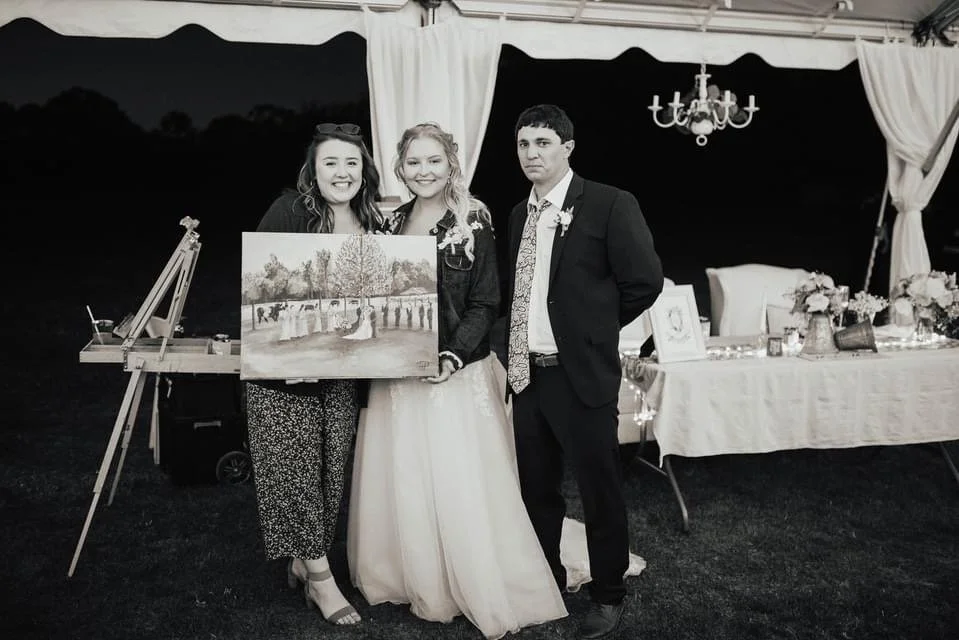 Three people standing together at a wedding reception, two women and one man, with the woman in the center wearing a wedding dress. The woman on the left is holding a painting, and there is a decorated table in the background with flowers and framed 