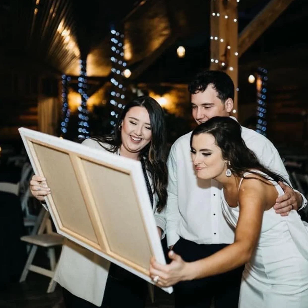 Three people, two women and one man, look happily at a picture frame at an indoor event with warm lighting and string lights in the background.