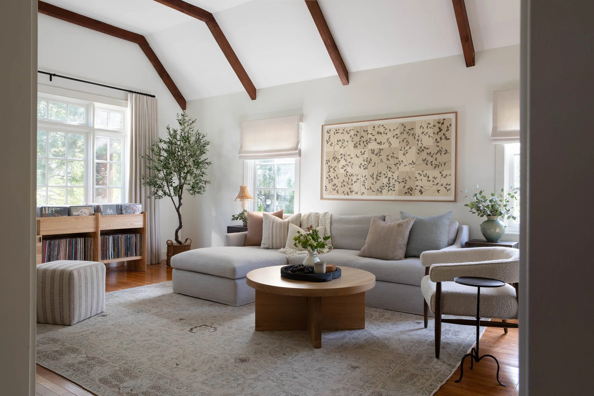 A cozy living room with white walls, wooden ceiling beams, large windows with cream curtains, a white sectional sofa with cushions, a wooden coffee table with decorative items, a floor lamp with a wicker shade, a bookshelf with records, a potted plant, and framed artwork on the wall.