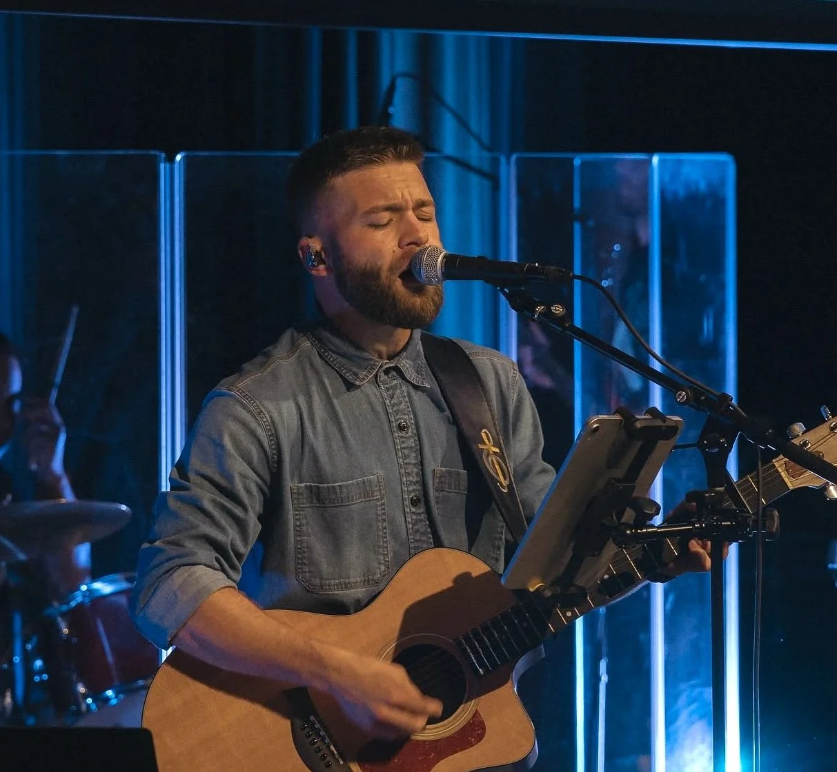A man with a beard and short hair, wearing a denim shirt, is singing into a microphone while playing an acoustic guitar during a live performance. He appears to be deeply immersed in the music.