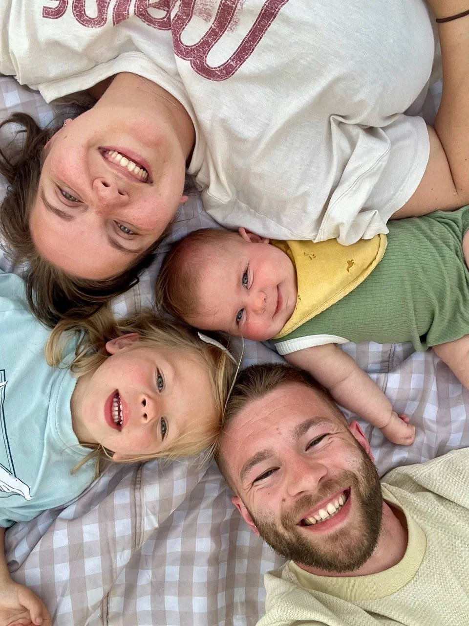 A family of four lying on a bed, smiling. The mother, father, and two children, a girl and a baby boy, are all smiling and looking at the camera.