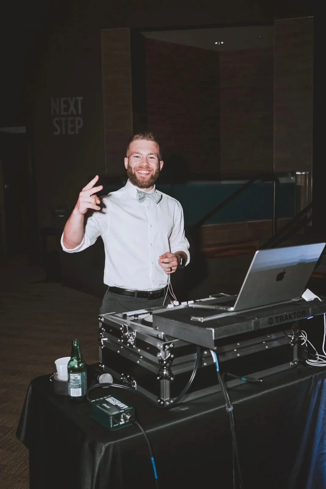 A man with a beard and mustache, wearing a white shirt and a bow tie, smiling and making a peace sign with his right hand, standing behind a DJ setup with a laptop and sound equipment in a dimly lit room.