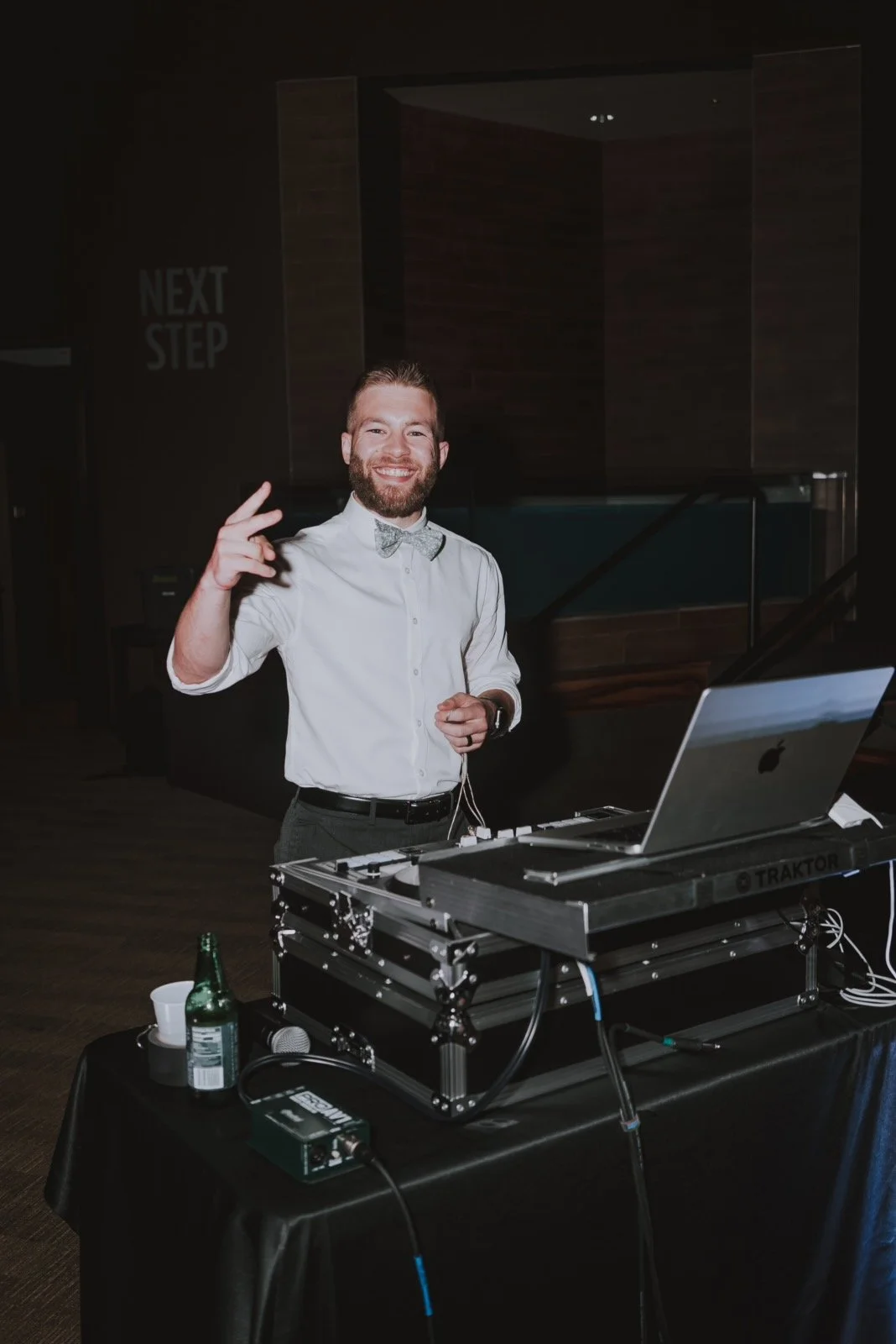 A smiling man with a beard, wearing a white shirt and bow tie, standing behind DJ equipment with a laptop, in a dimly lit indoor setting. He is making a peace sign with his right hand.