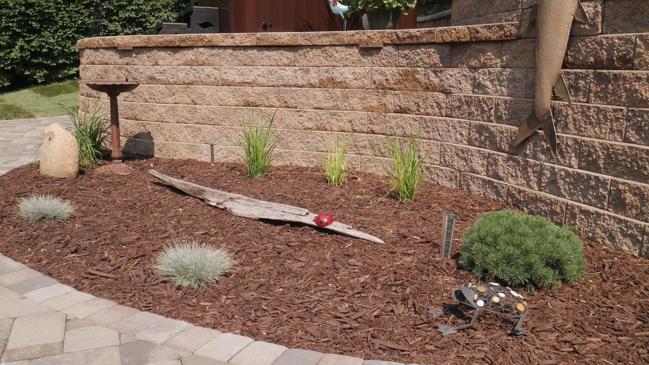 Garden bed with brown mulch, small green shrubs, and decorative garden ornaments including a ladybug on a piece of driftwood, a frog statue with coins on its back, and a fish sculpture attached to the brick wall in the background.