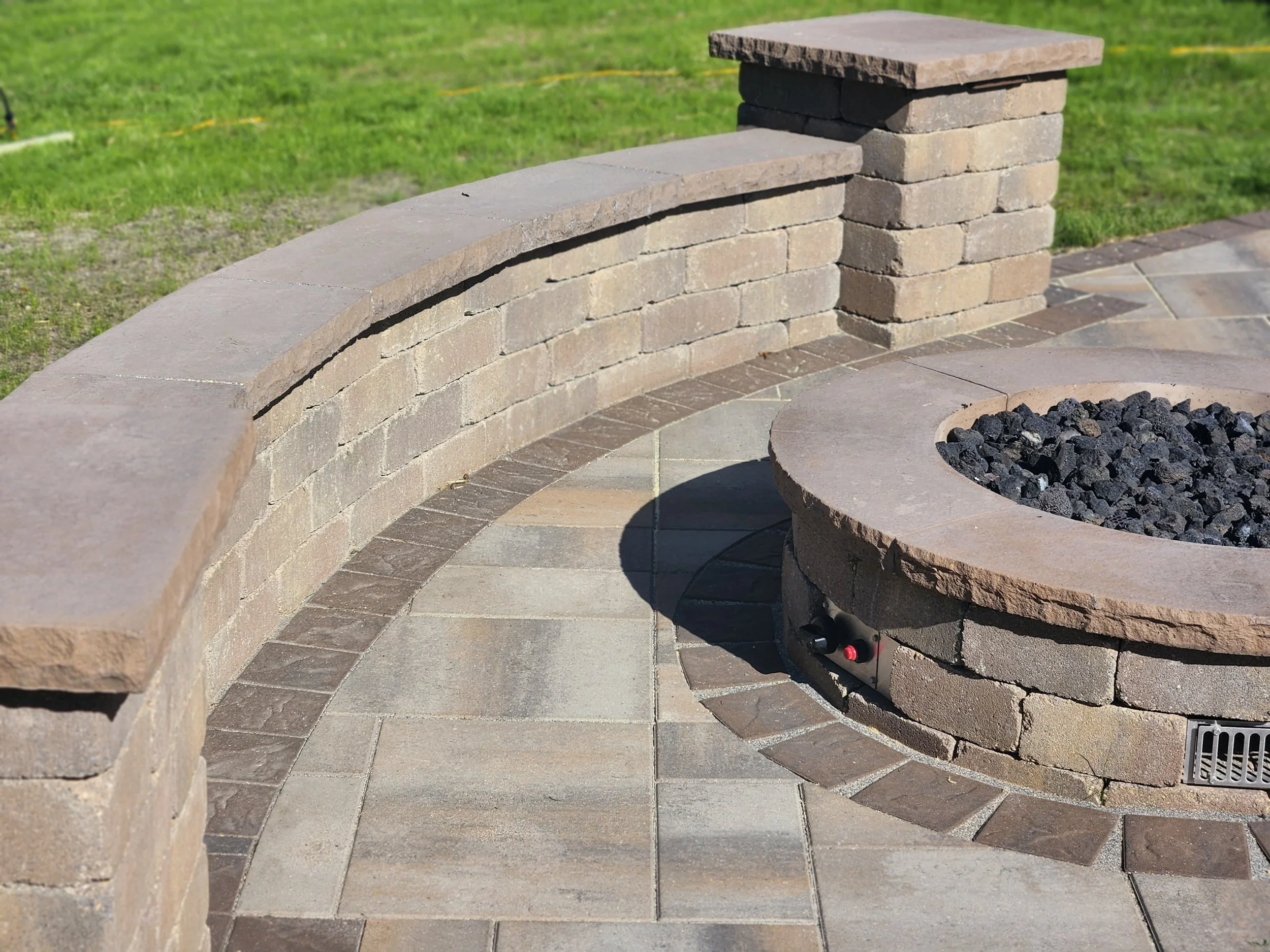 A circular stone fire pit surrounded by a curved low brick wall and paved patio with grass in the background.