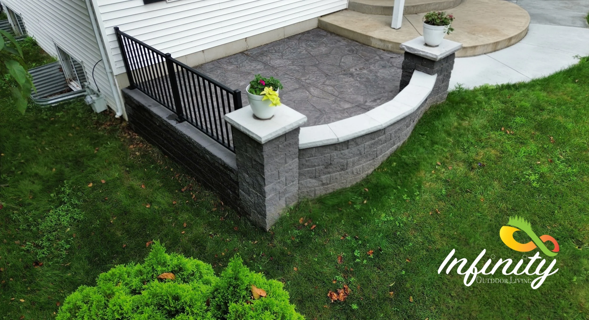 Backyard patio with stone and concrete finishing, black metal railing, and potted plants on the ledge, surrounded by green grass and bushes.