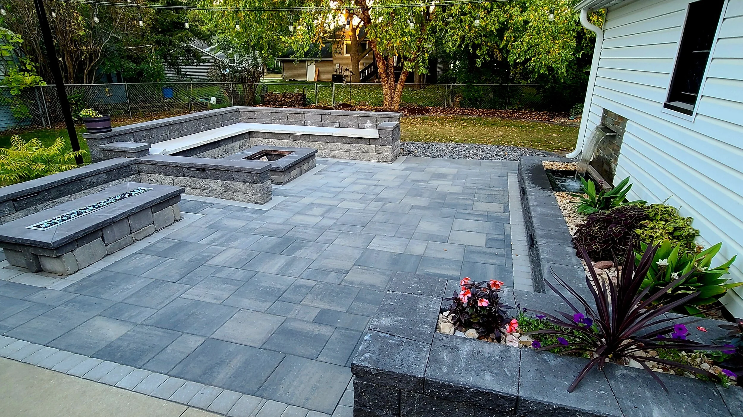 Backyard patio with stone pavers, built-in seating, a fire pit, and a garden bed with plants and flowers beside a white house.