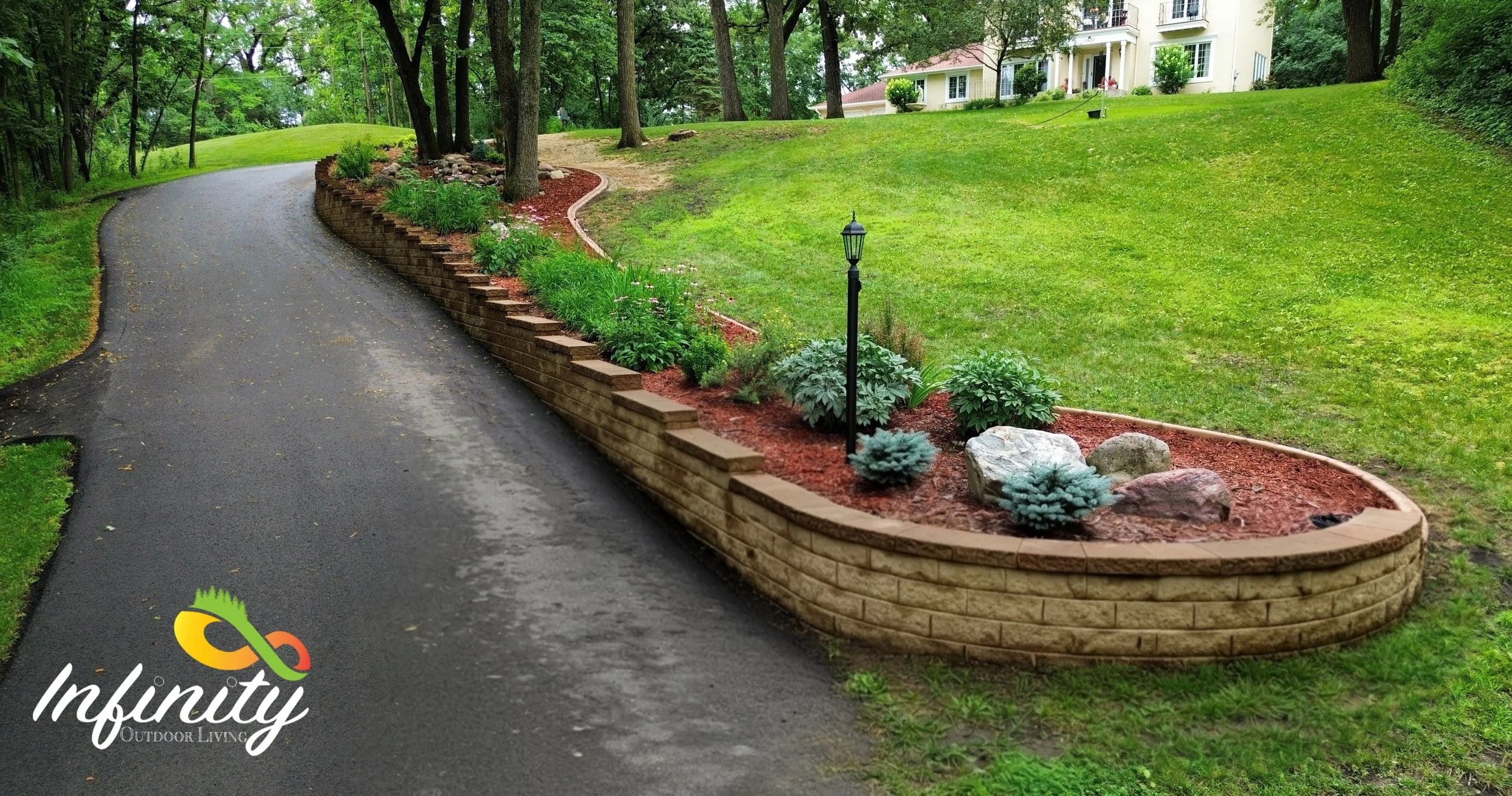 A curved driveway runs past a landscaped garden bed with small shrubs, flowers,  and rocks, bordered by a wooden retaining wall, in front of a yellow house surrounded by trees.