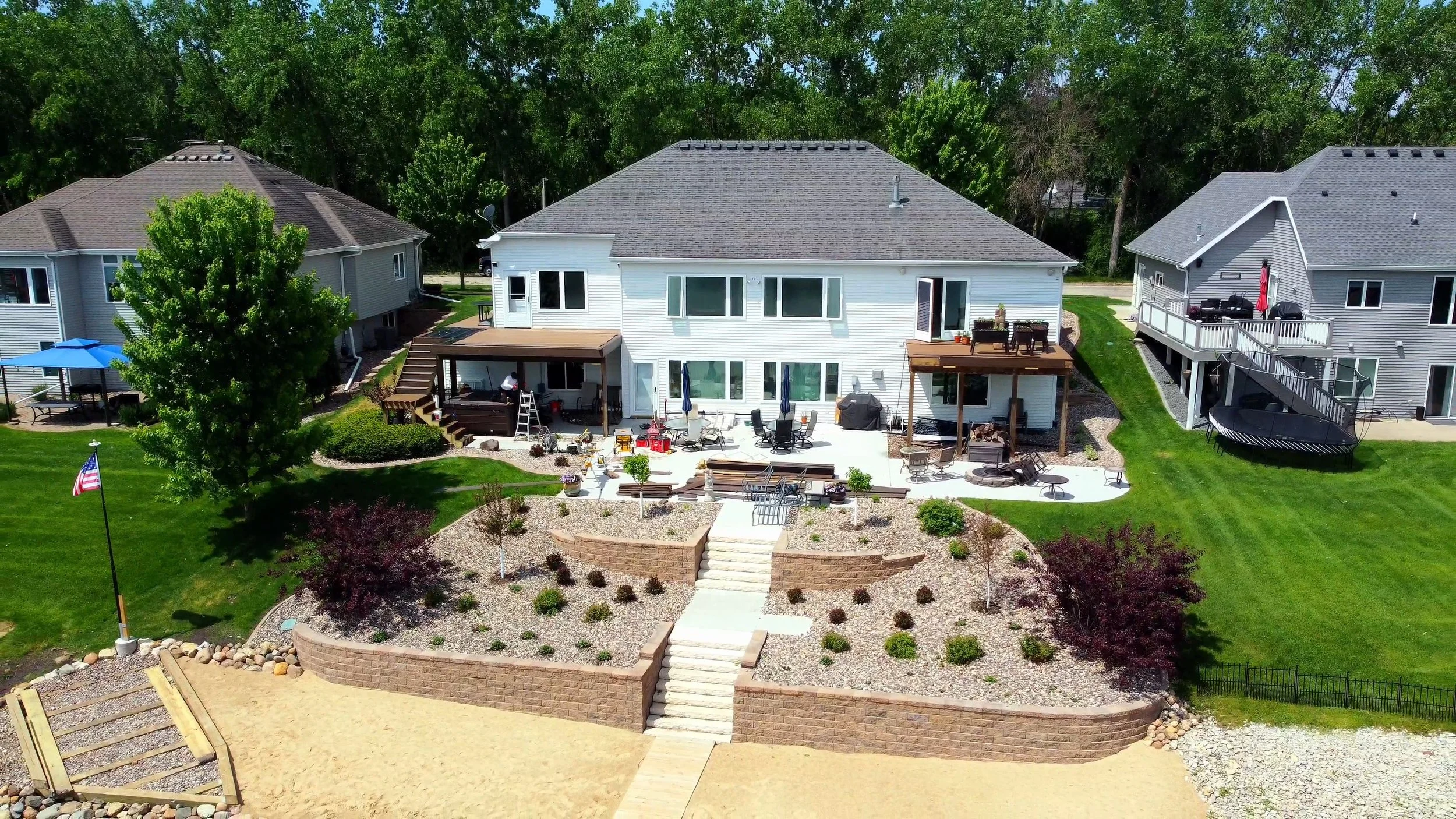 Aerial view of a large backyard of a white house with multiple decks, surrounded by green grass, trees, and neighboring houses.