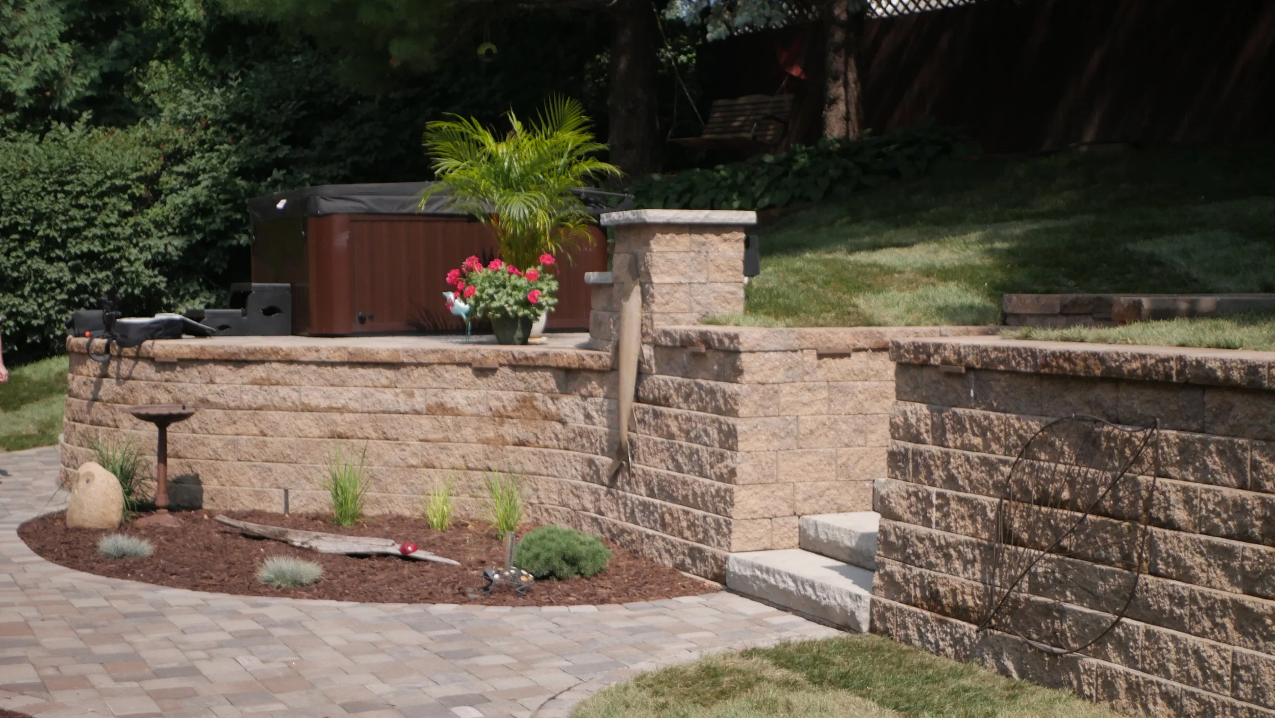 Backyard with brick retaining wall, hot tub, potted plants, garden decorations, and a paved patio area.