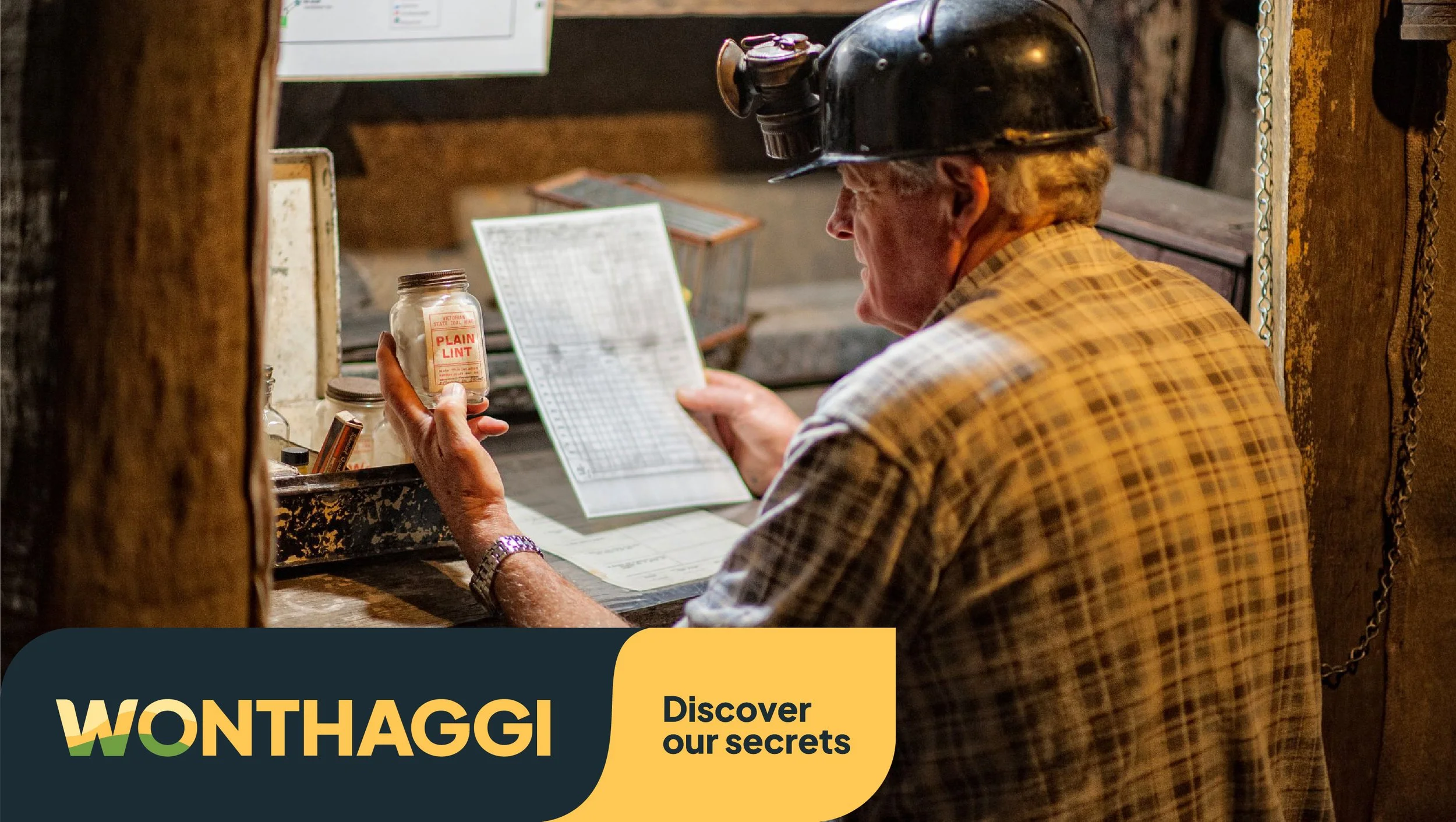 Older man visits mining museum experience, examining infosheet and old jar of lint while wearing old helmet