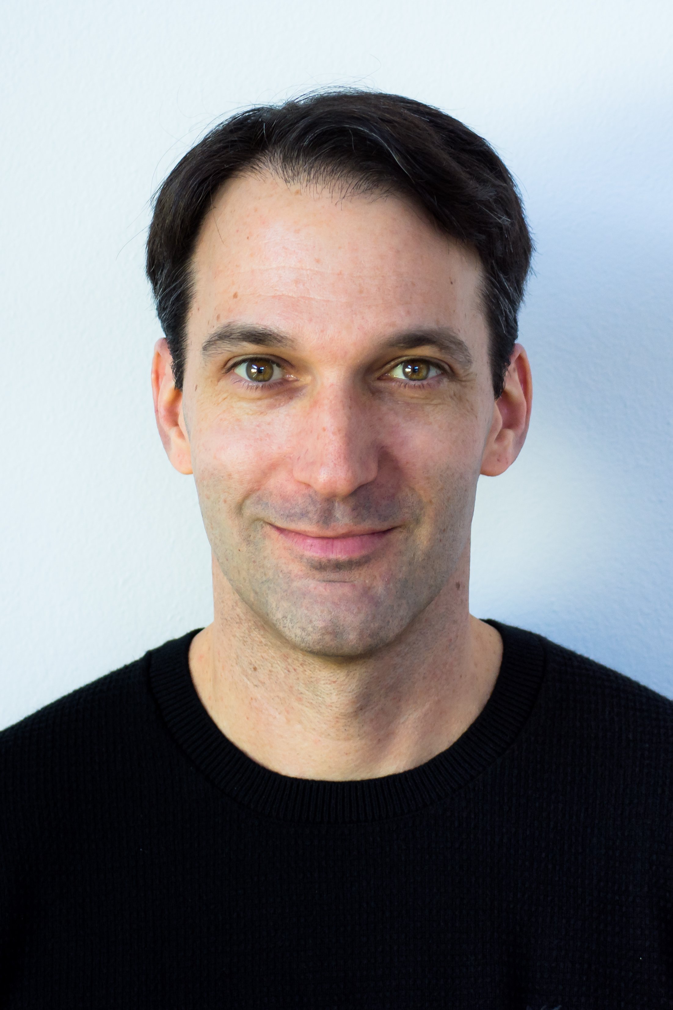 Portrait of a man with dark hair, hazel eyes, slight smile, wearing a black sweater, against a plain light background.
