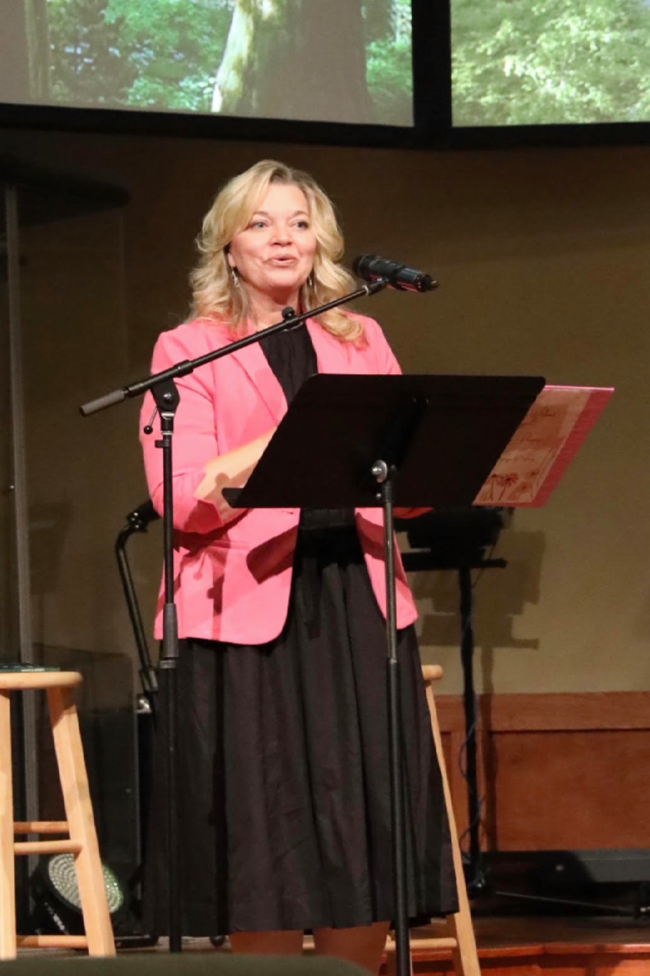 A woman with blonde hair wearing a pink blazer and black dress stands behind a music stand, speaking into a microphone, with a screen displaying a forest scene in the background.
