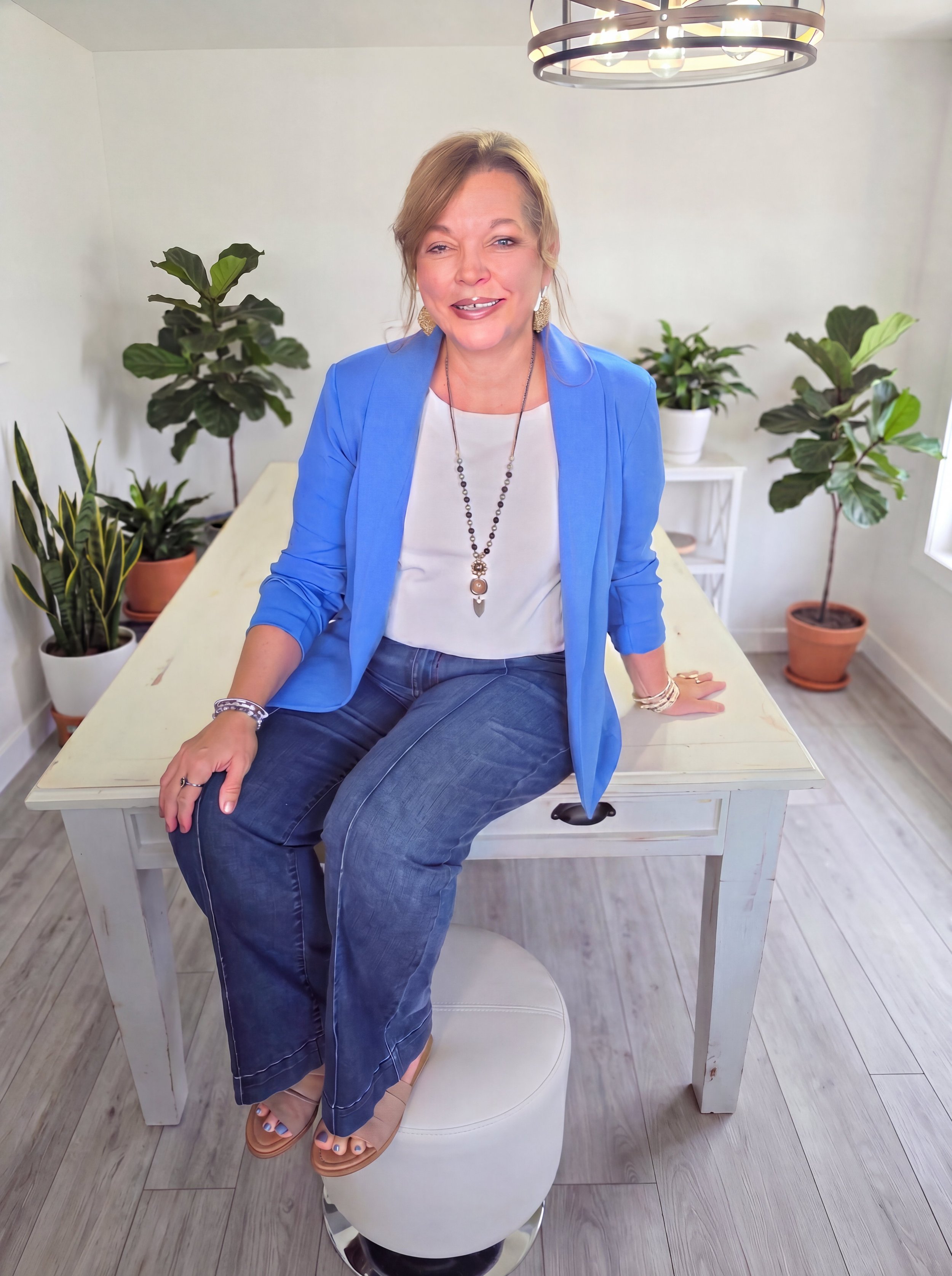 A woman with blonde hair smiling, wearing a blue blazer, white top, and jeans, sitting on a white stool in a room with potted plants, a white table, and a black circular chandelier.