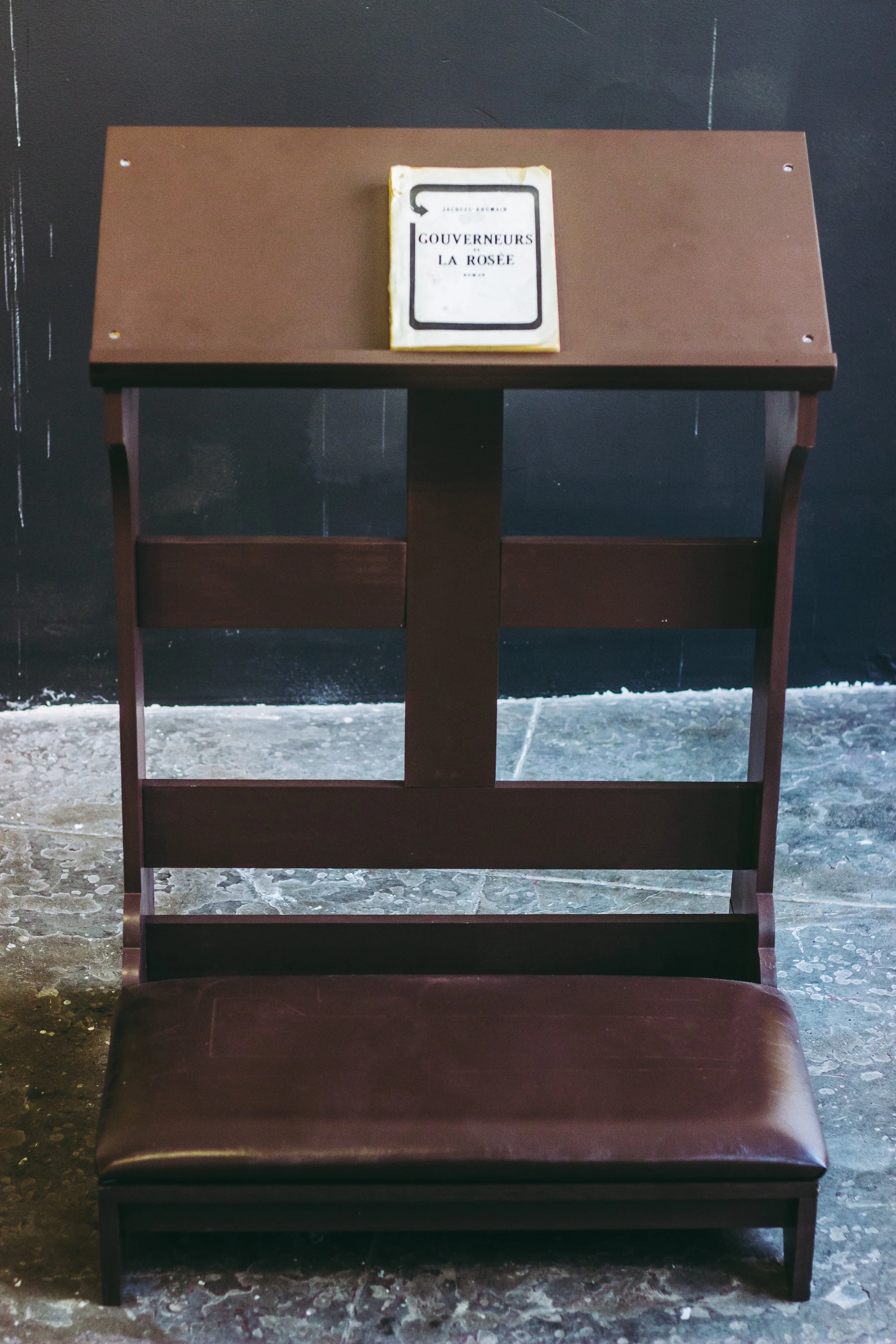 The kneeler placed before the photographs invites viewers into an act of collective reflection. While kneeling traditionally directs one's gaze upward toward heaven and God, this installation instead asks viewers to look up to community. In Masters o