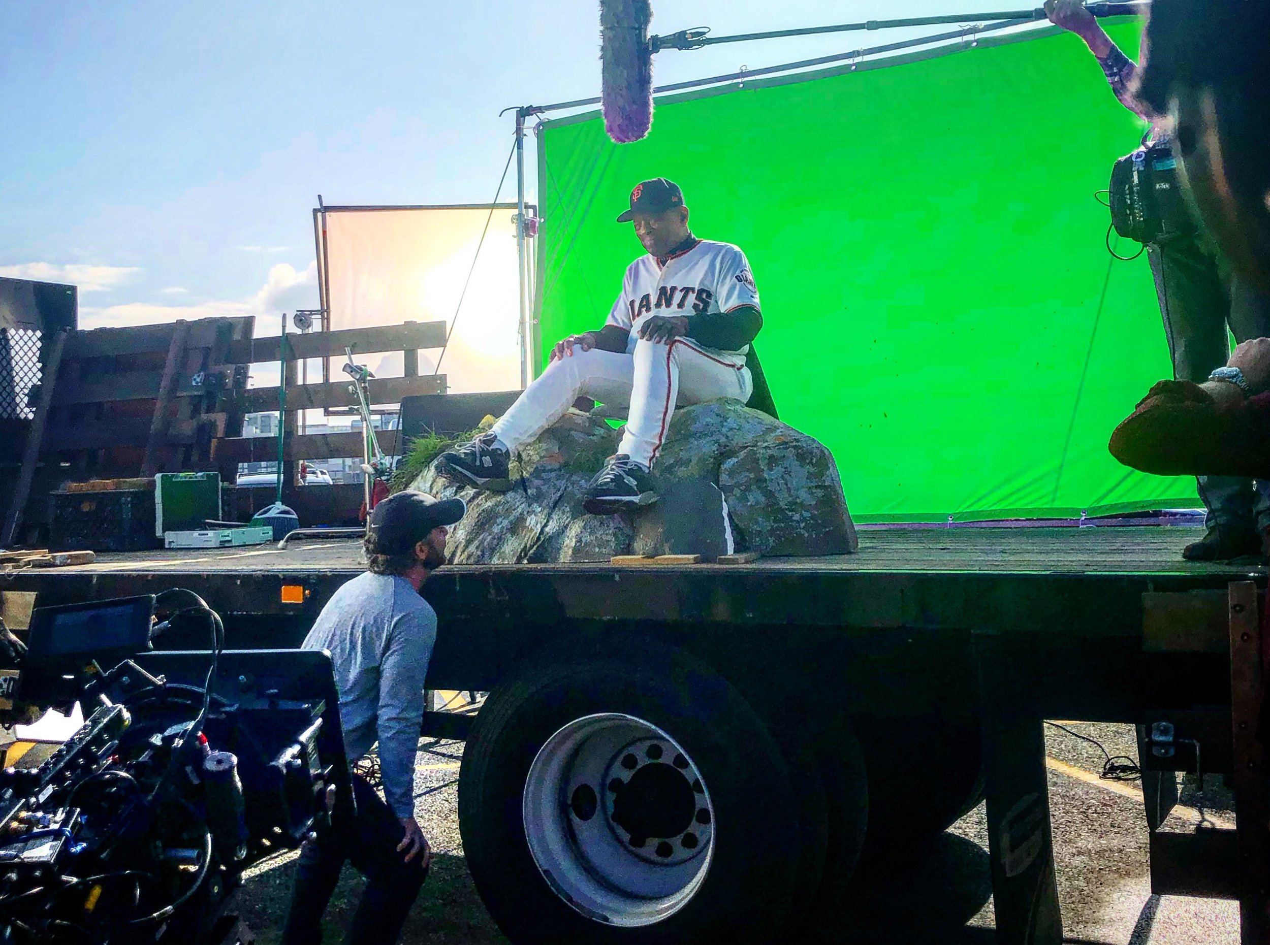 Behind the scenes of a Toyota commercial shoot with Willie Mays in a San Francisco Giants baseball uniform sitting on a large rock, green screen in background, crew members and equipment around.