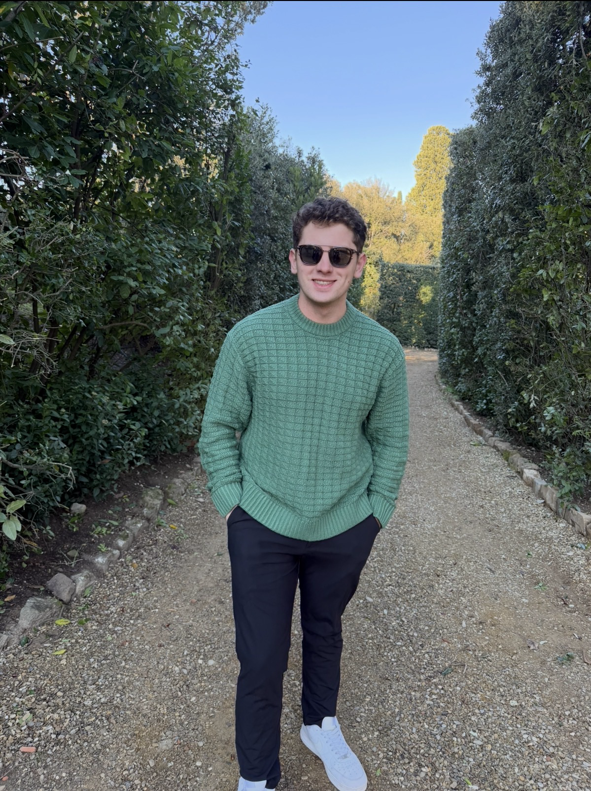 A young man with curly hair and sunglasses, wearing a green sweater, black pants, and white shoes, standing on a gravel path surrounded by green bushes and trees under a clear blue sky.