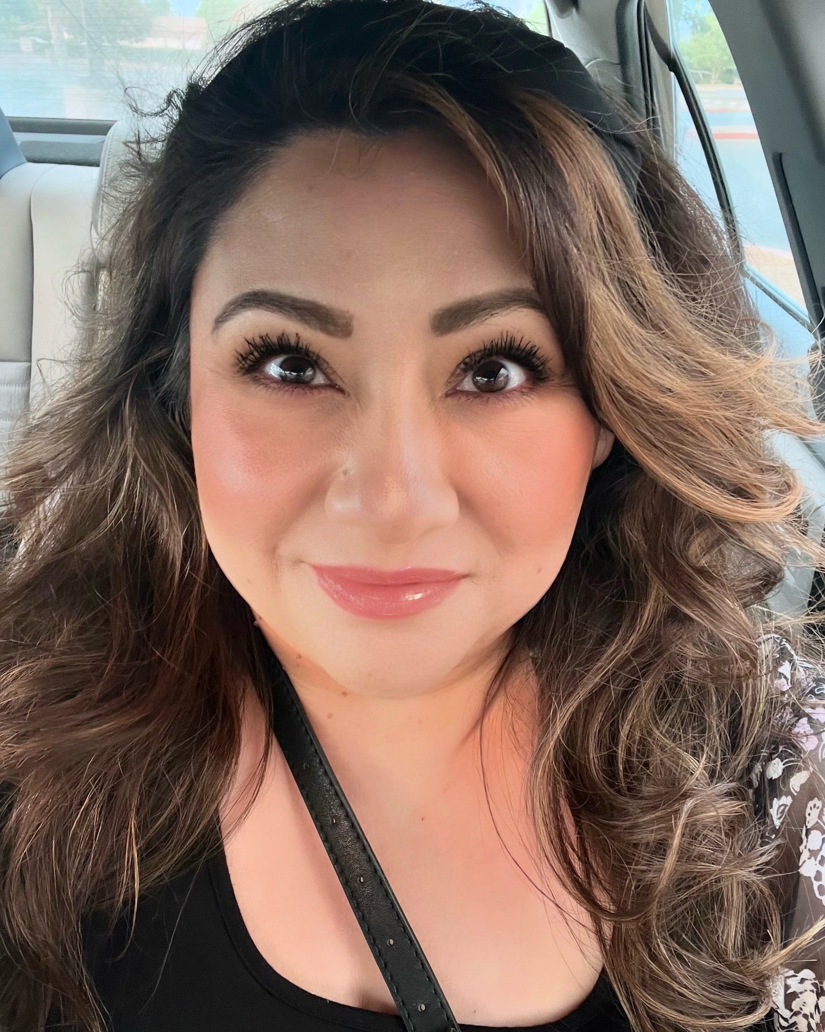 A smiling woman with dark brown wavy hair, wearing makeup, inside a vehicle with a view of a parking lot and buildings through the window.
