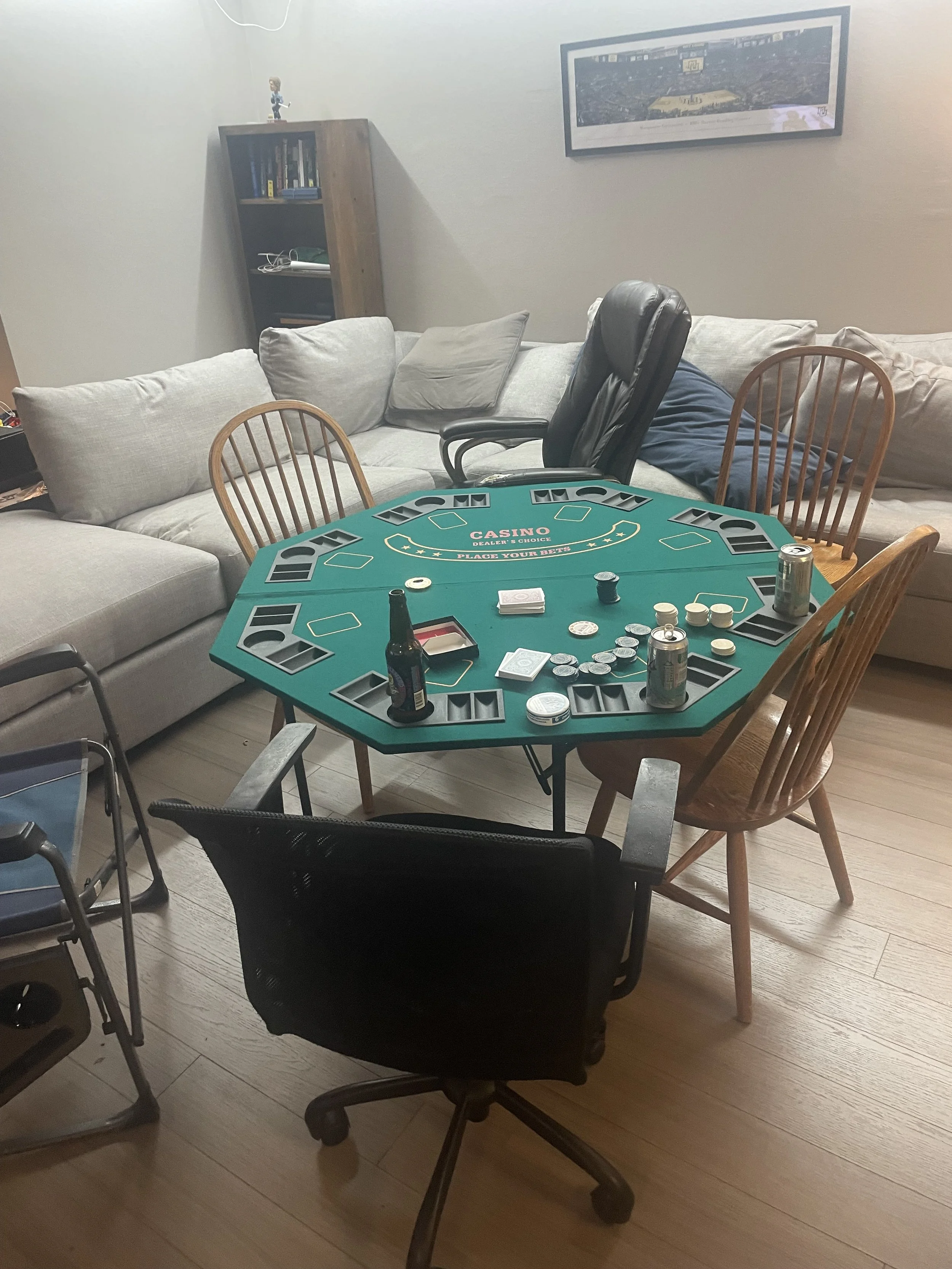 A group of tables in a room set up for poker, with poker chips, bottles, and cans on top. Surrounding the tables are various chairs, including office chairs and wooden dining chairs. In the background, there is a beige sectional sofa with cushions, a