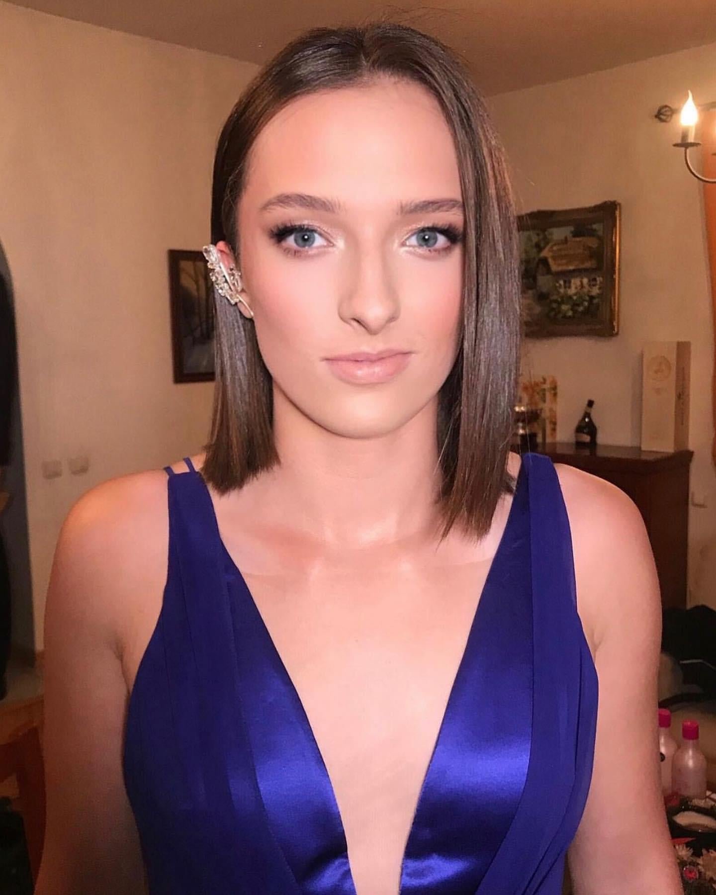 A young woman with straight brown hair and blue eyes wearing a royal blue dress and a decorative earring, standing indoors with a neutral background.
