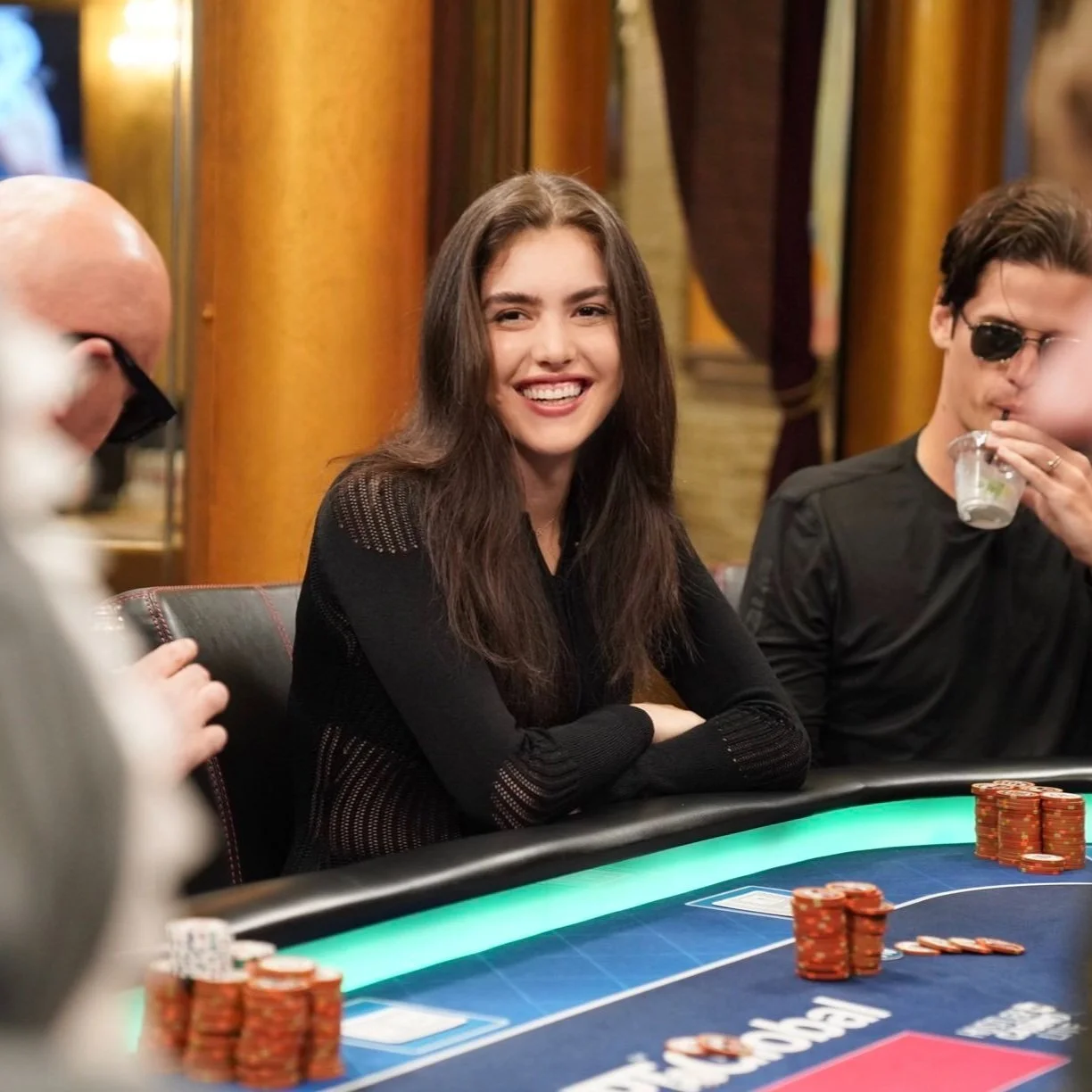 A young woman with long brown hair smiling at a poker table with poker chips, surrounded by other players in a casino setting.
