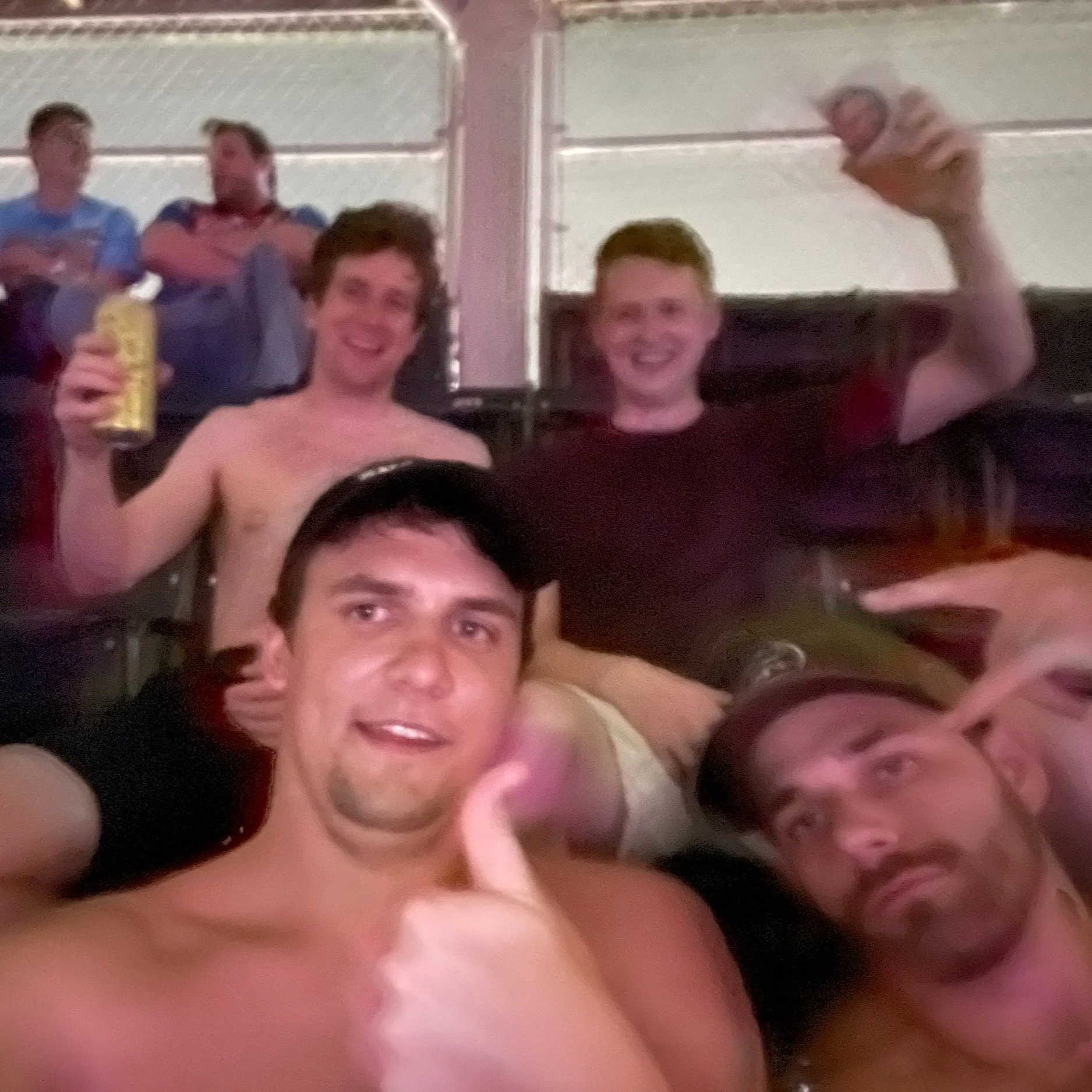 Group of men at a party, some shirtless, smiling and making gestures, in a casual indoor setting with other people in the background.