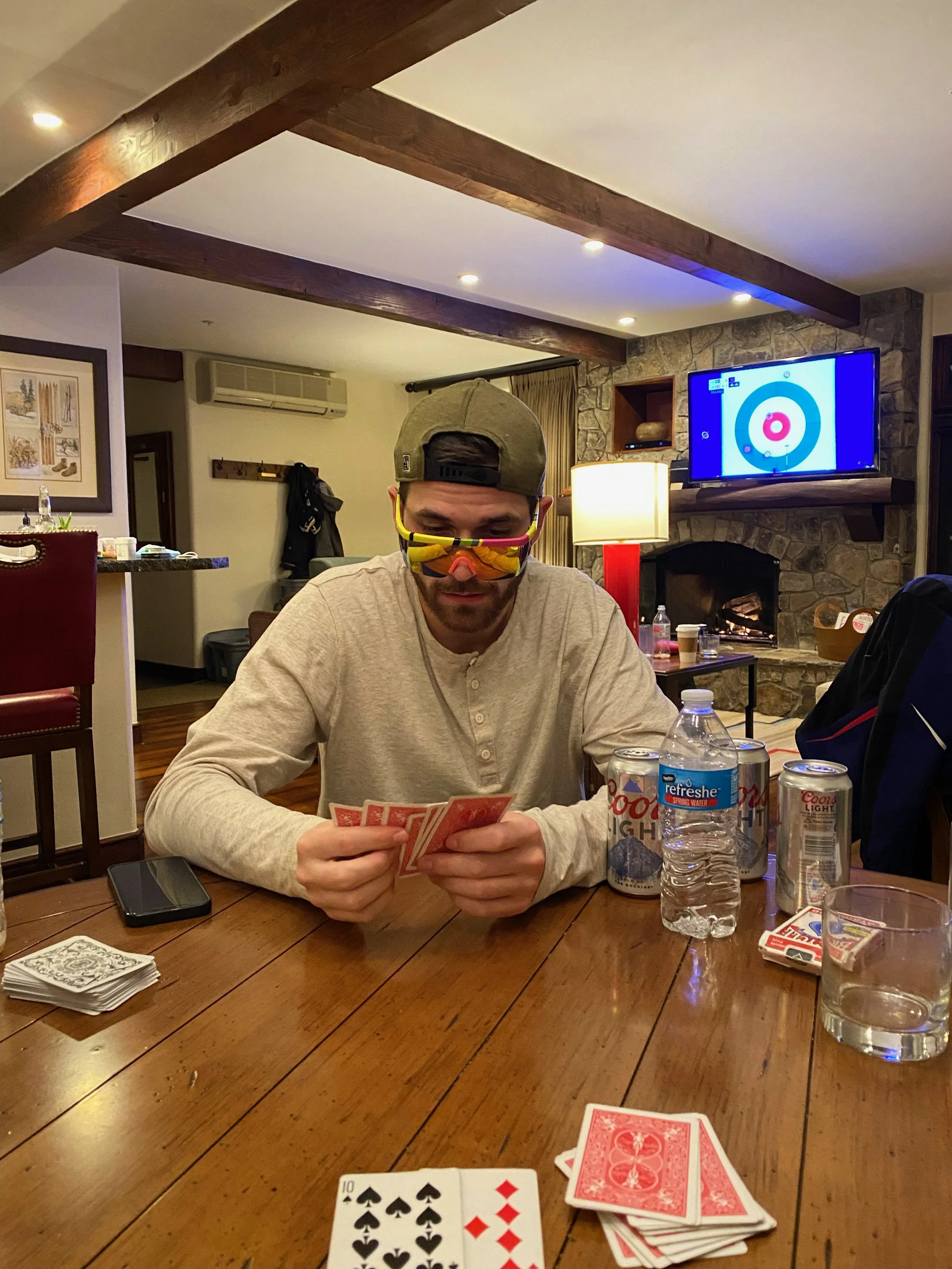 A man sitting at a wooden table playing cards, wearing sunglasses, a baseball cap, and a beige long-sleeve shirt, with drinks and playing cards on the table, in a cozy room with a TV mounted on a stone fireplace.