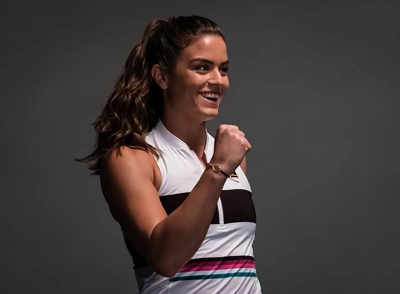 A young woman smiling and posing with her fist raised, wearing a sleeveless athletic top with black, white, pink, and teal stripes, and a bracelet on her wrist, against a gray background.