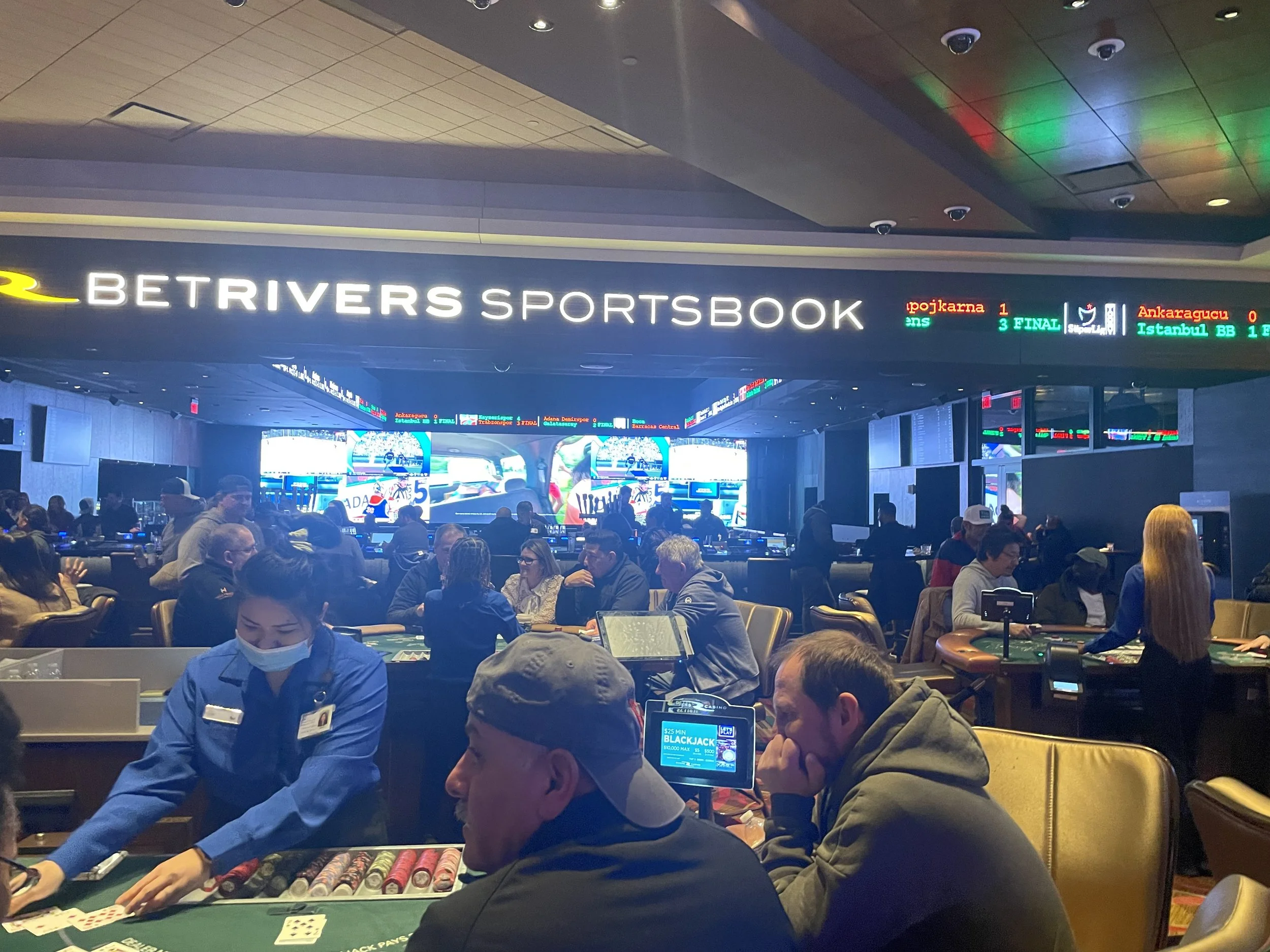 People playing poker at a gambling table in a casino with a large digital sign reading 'BETRIVERS SPORTSBOOK' overhead. There are multiple screens and electronic displays around the casino, showing sports scores and betting odds.