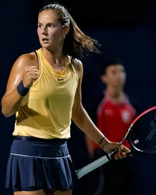 A young woman in a yellow sports top and dark skirt holding a tennis racket on a tennis court.