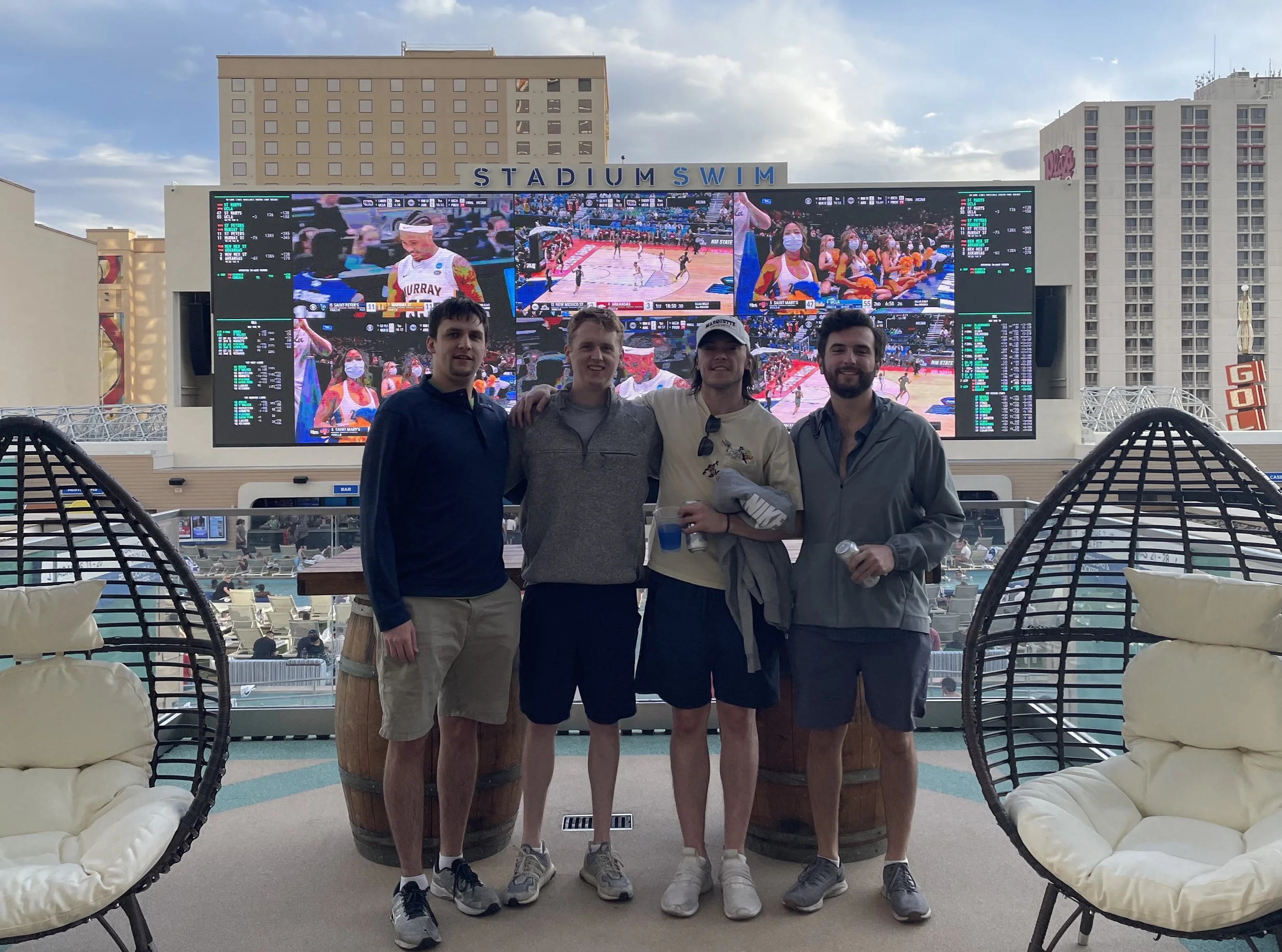 Four men standing together at an outdoor area with lounge chairs, with a large digital screen behind them displaying a live basketball game. The background shows buildings and an overcast sky.