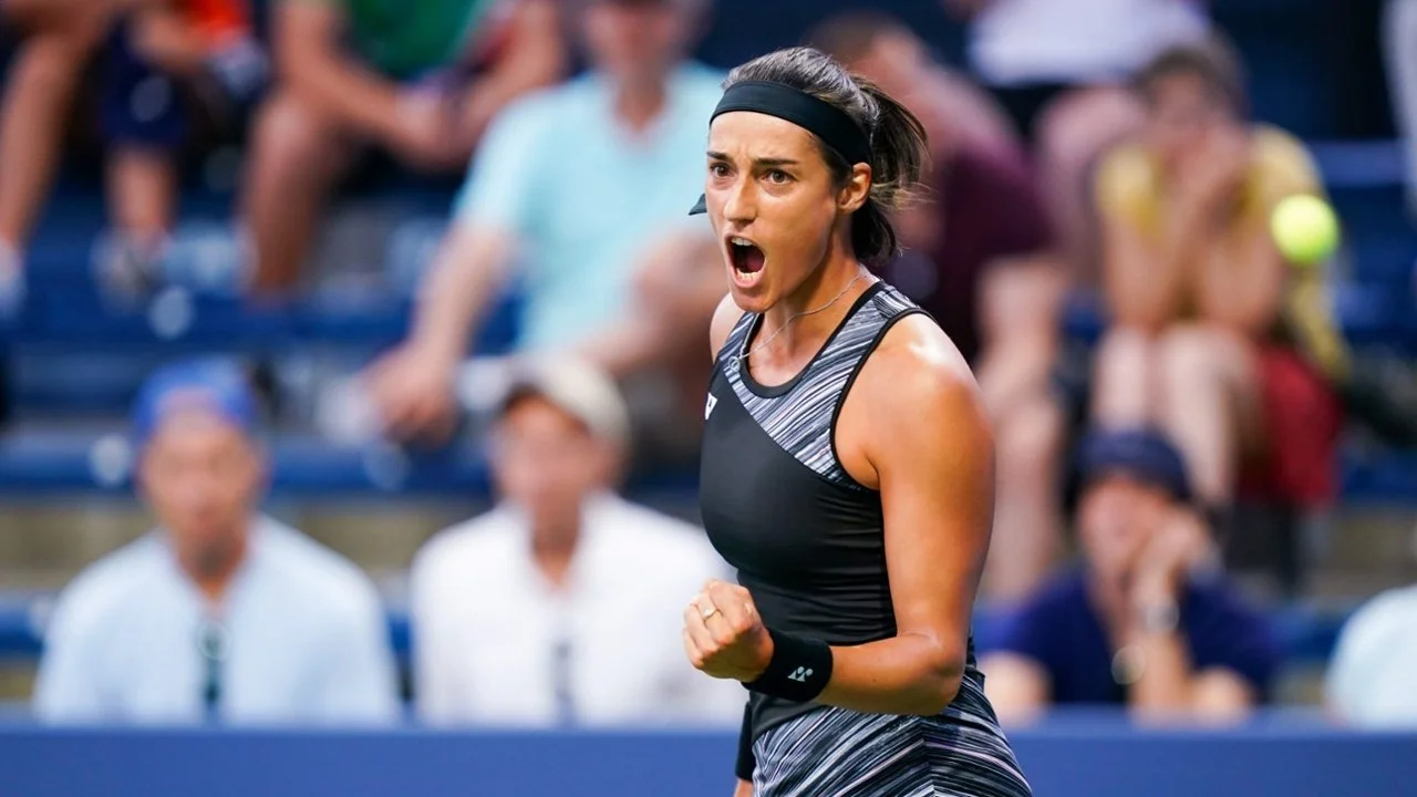 Tennis player celebrating on the court with a clenched fist and an expression of triumph, wearing a black athletic outfit and headband, with spectators in the background.