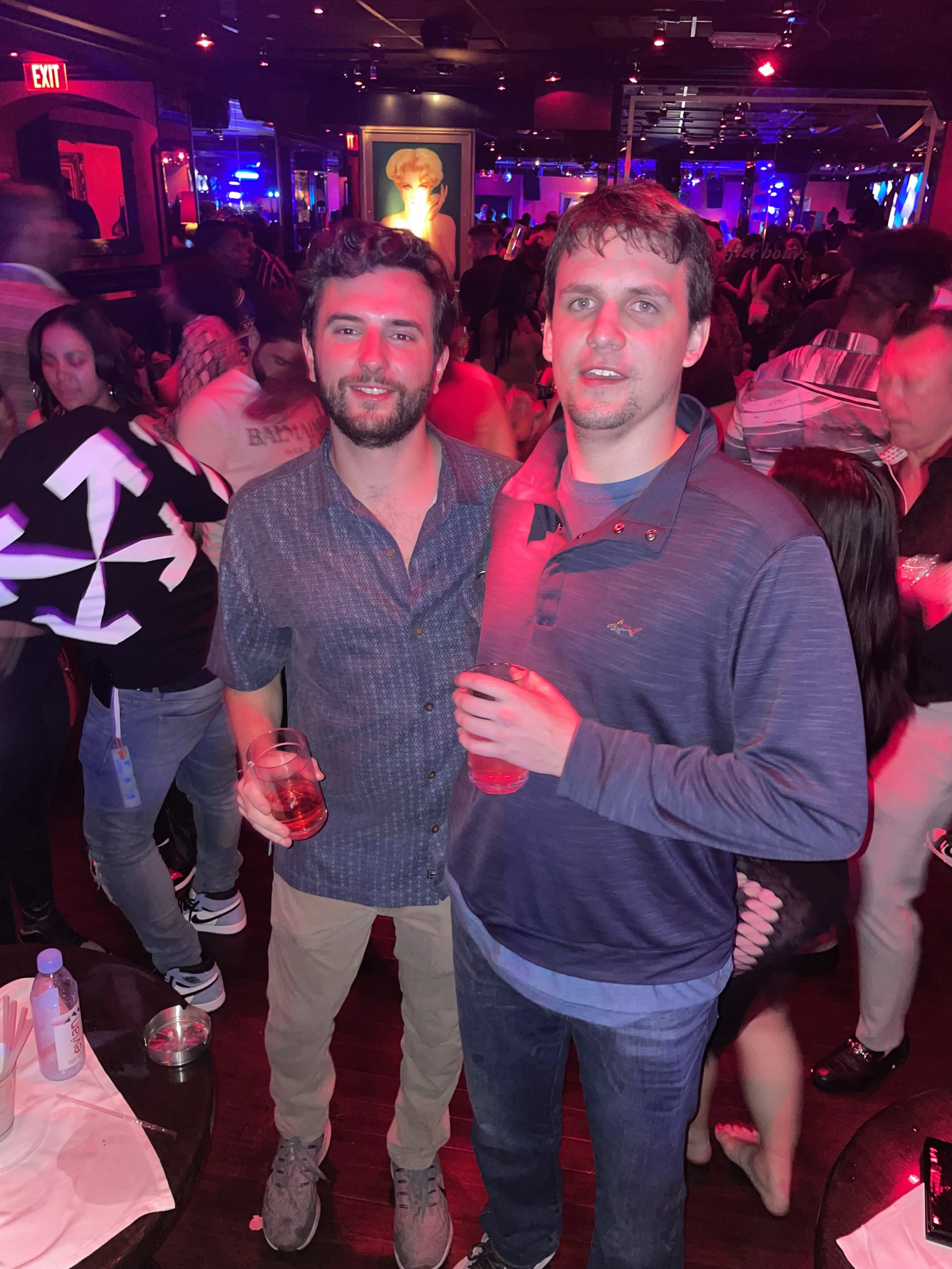 Two men with drinks at a crowded nightclub with colorful lights and artwork on the walls.