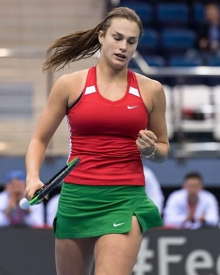 Female tennis player in a red tank top and green skirt on a tennis court.