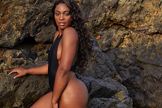 A woman with long curly hair sitting on rocks near water, wearing a black swimsuit.