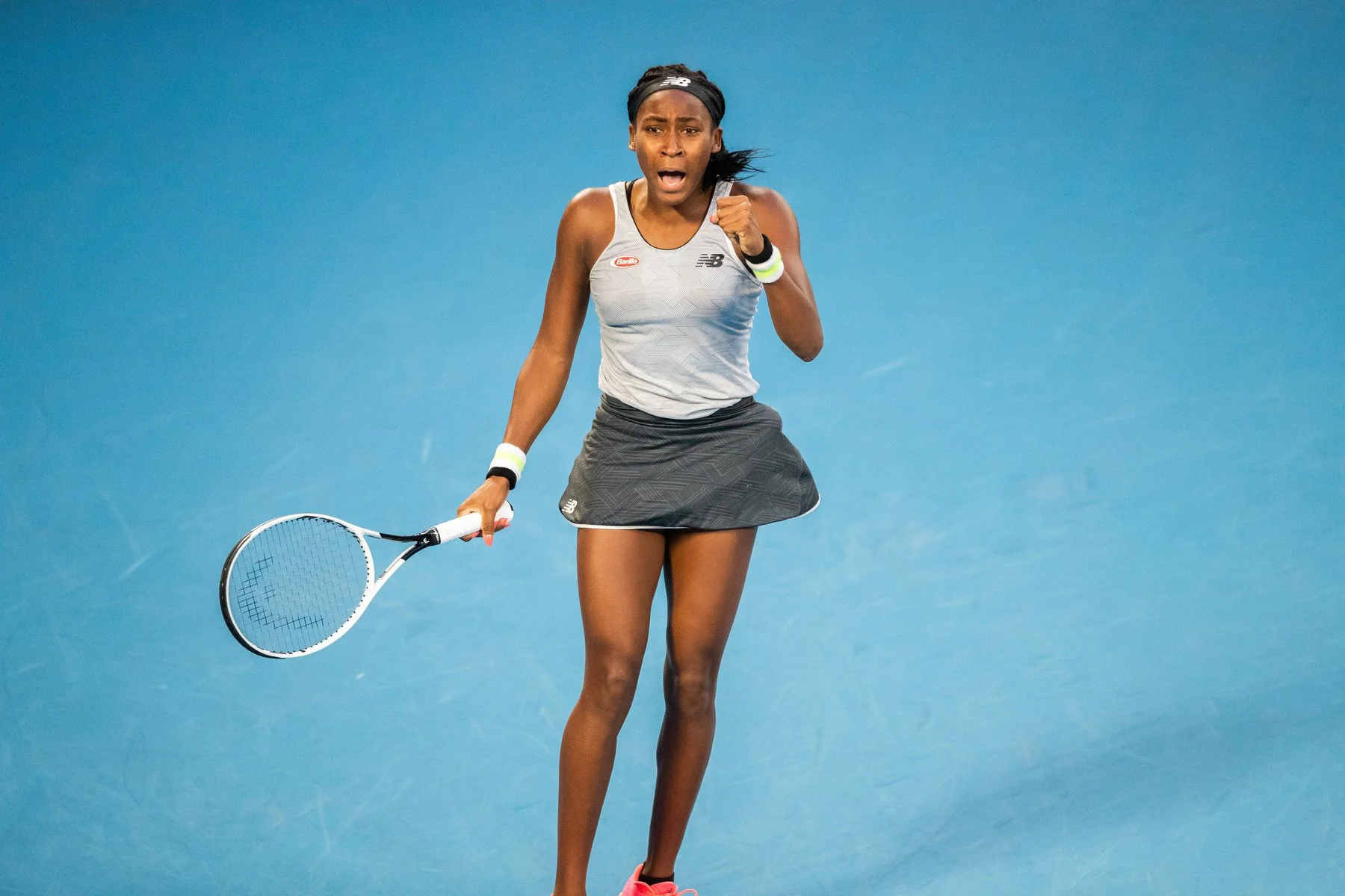 A female tennis player on a blue court displays a surprised or intense expression, holding a tennis racket in her right hand, wearing a gray athletic top, a black skirt, and pink shoes.