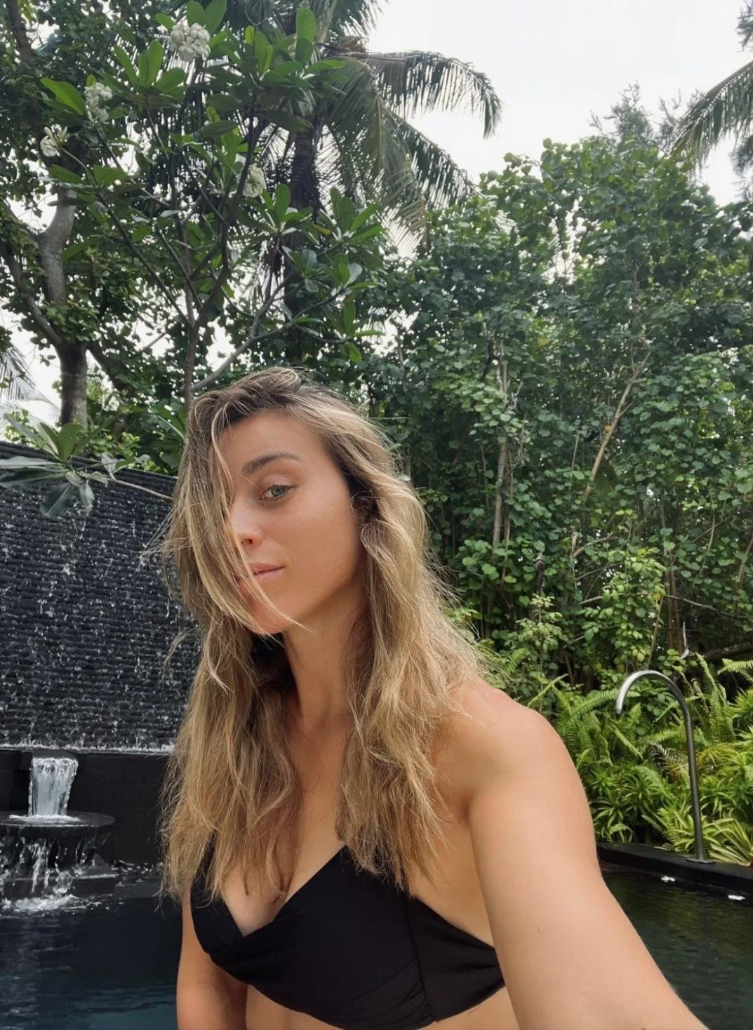 A woman taking a selfie outdoors near a water fountain, surrounded by lush green tropical plants and trees, with overcast sky.
