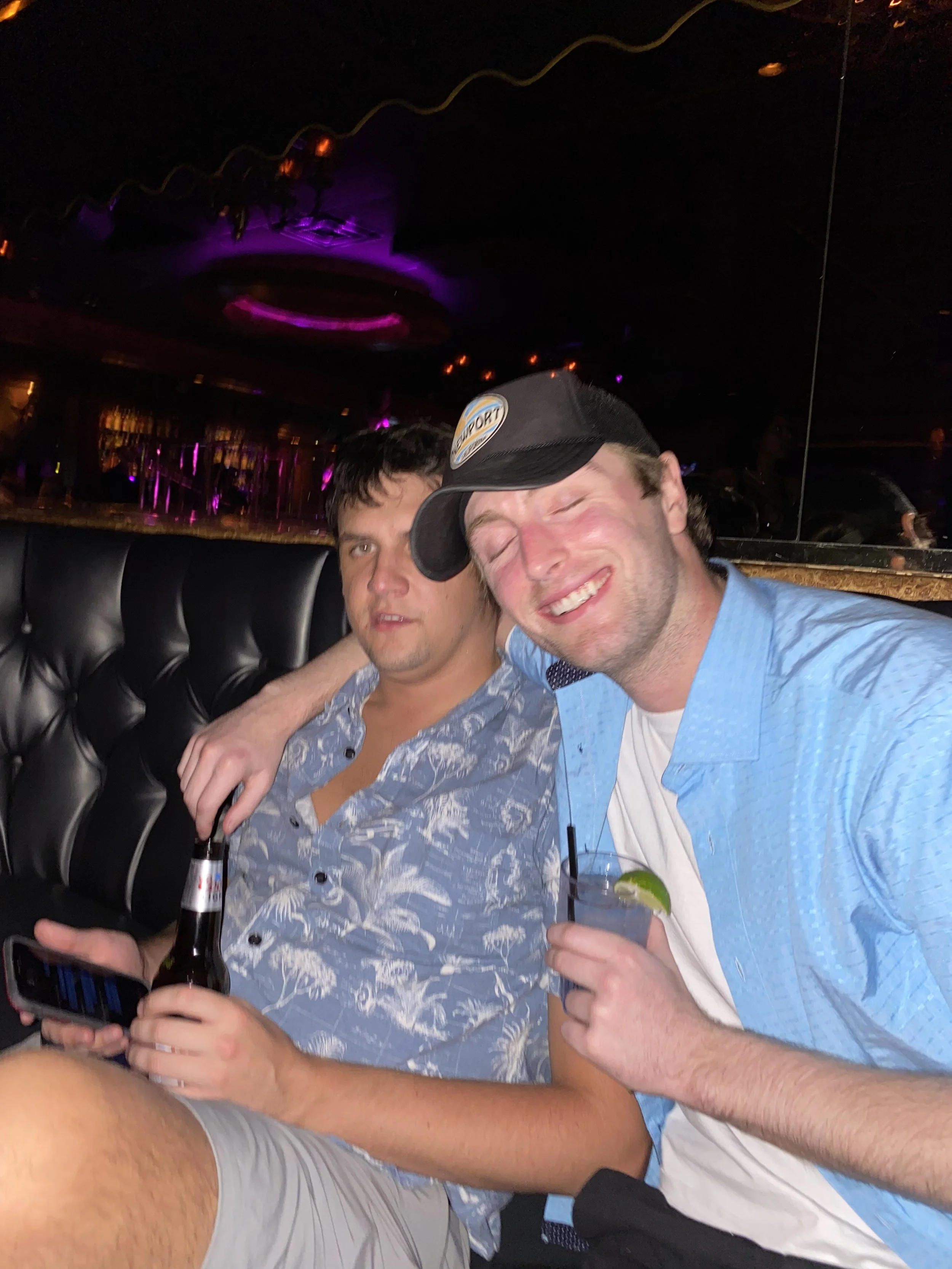 Two men sitting together in a nightclub, one holding a beer and the other holding a drink with lime, smiling and leaning into each other, with a colorful, illuminated background.