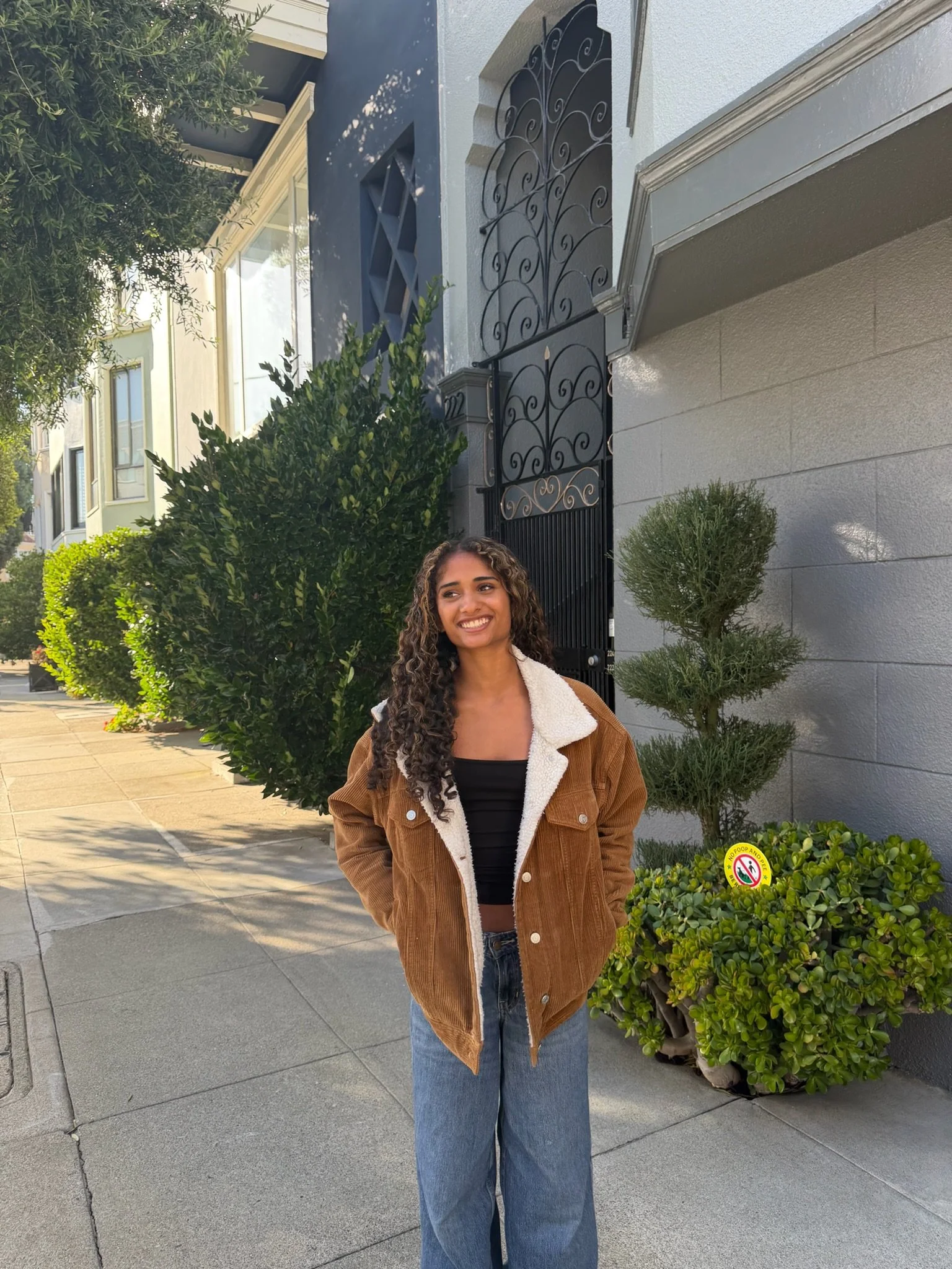 Young woman sitting on a curved outdoor wall, wearing a black top, denim shorts, and black sneakers with white laces, smiling at the camera with cars and trees in the background.