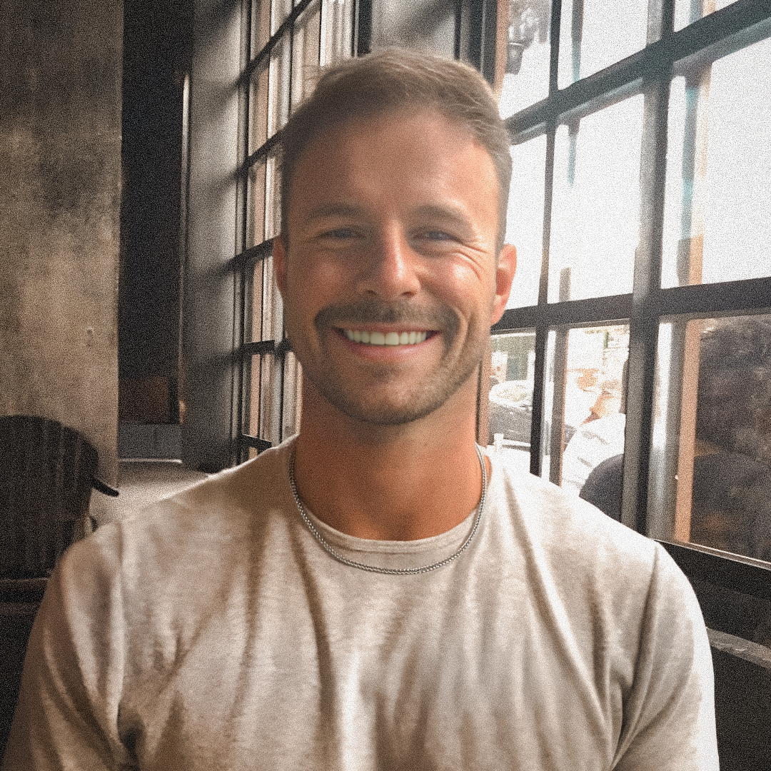 A smiling man with short light brown hair, wearing a light gray t-shirt and two silver necklaces, sitting in front of a large window with multiple panes inside a rustic-style cafe.