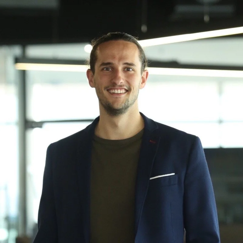 Smiling man in a dark blazer and t-shirt standing indoors by windows