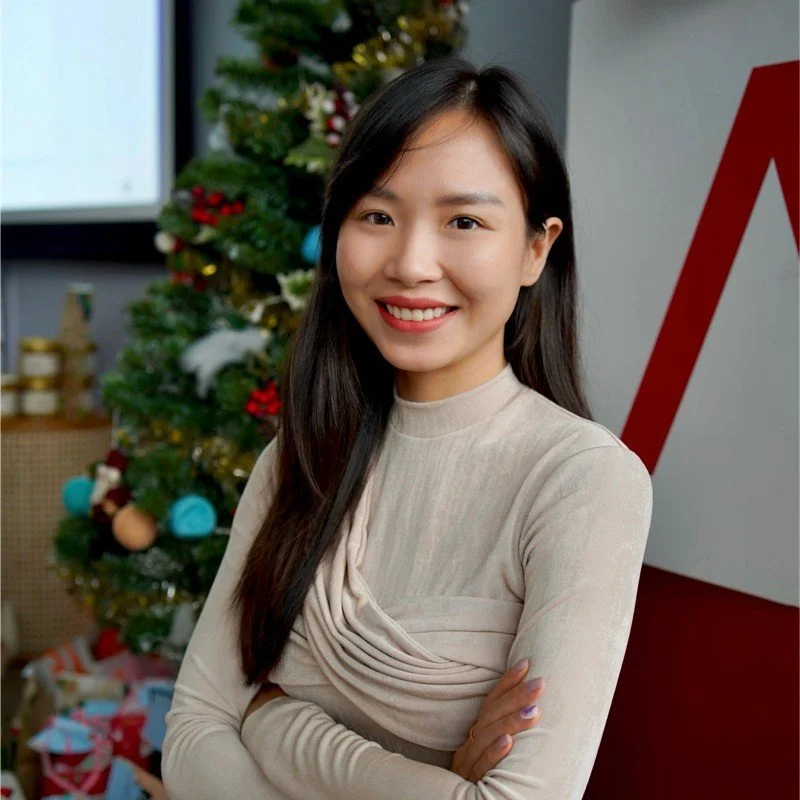 A smiling woman with long dark hair standing in front of a decorated Christmas tree with colorful ornaments, indoor setting.