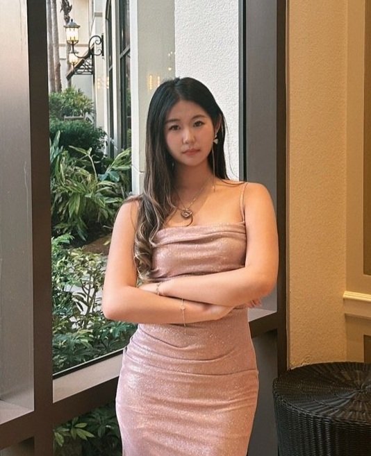 Young woman with long dark hair wearing a shiny pink dress, standing indoors next to a window with greenery outside.