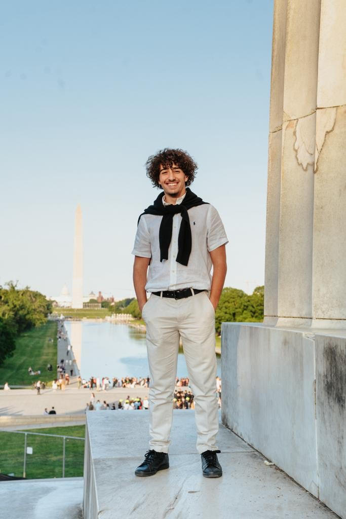 A young man with curly hair smiling, standing on a marble platform near the Washington Monument with the Reflecting Pool in the background, in Washington D.C.