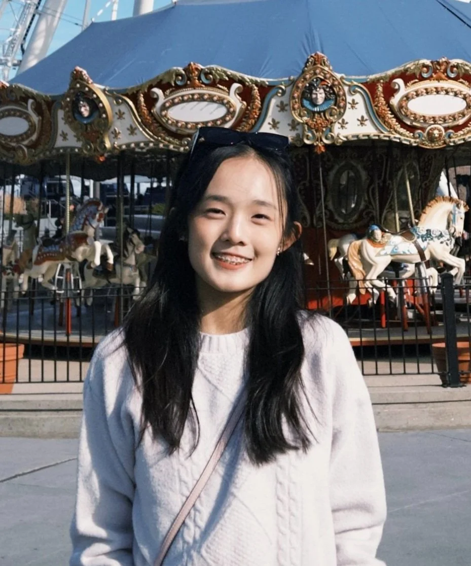 Young woman smiling in front of a vintage carousel with painted horses at an amusement park.