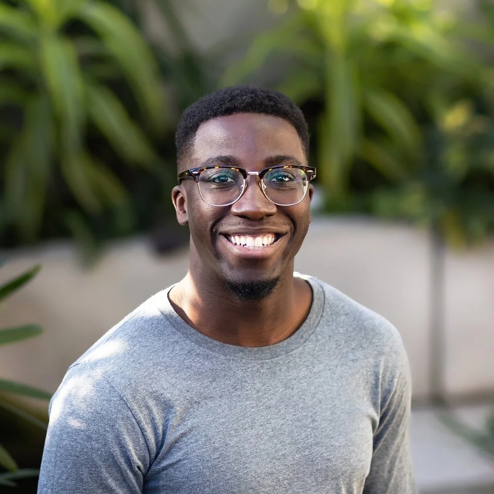 A smiling young man with glasses, wearing a gray shirt, standing outdoors with green foliage in the background.