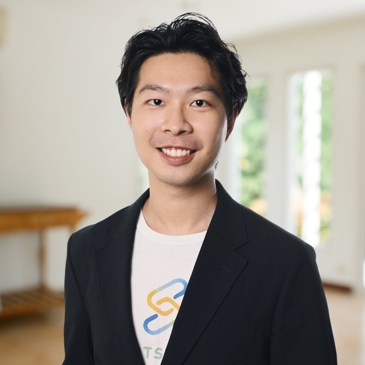 A young man with black hair smiles at the camera in an indoor setting with windows and greenery in the background.