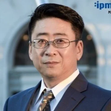 A man in a dark suit, wearing glasses, standing in front of a washing machine.