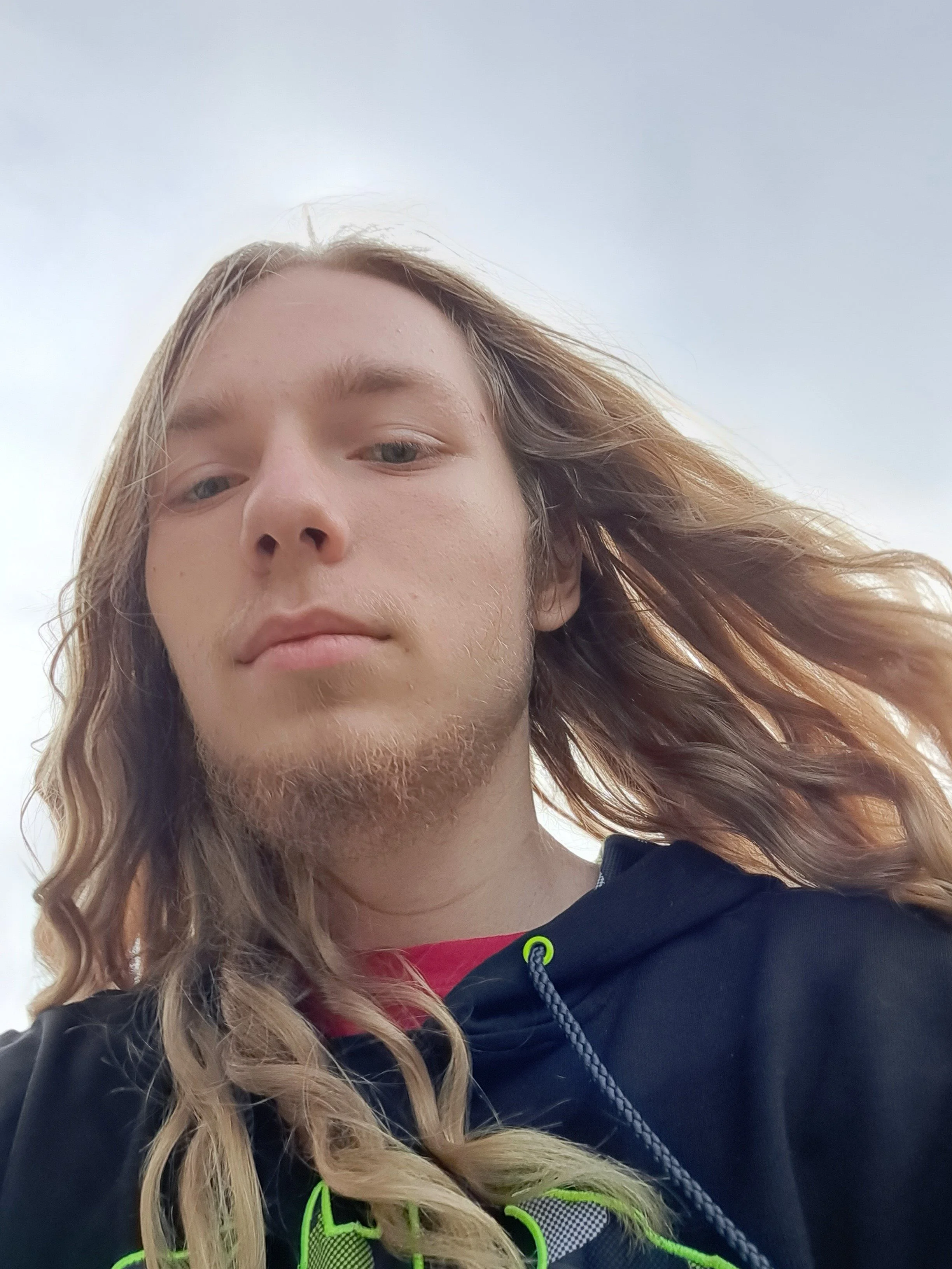 A close-up selfie of a young man with long, wavy blond hair, blue eyes, and a short beard. He is wearing a black hoodie with bright green accents and a red shirt underneath, outdoors against a cloudy sky.
