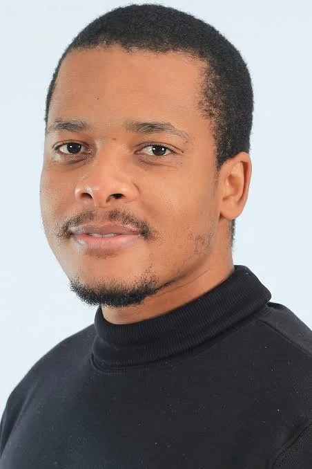 Portrait of a man with short dark hair, light skin, and a slight beard, wearing a black turtleneck sweater, against a light blue background.